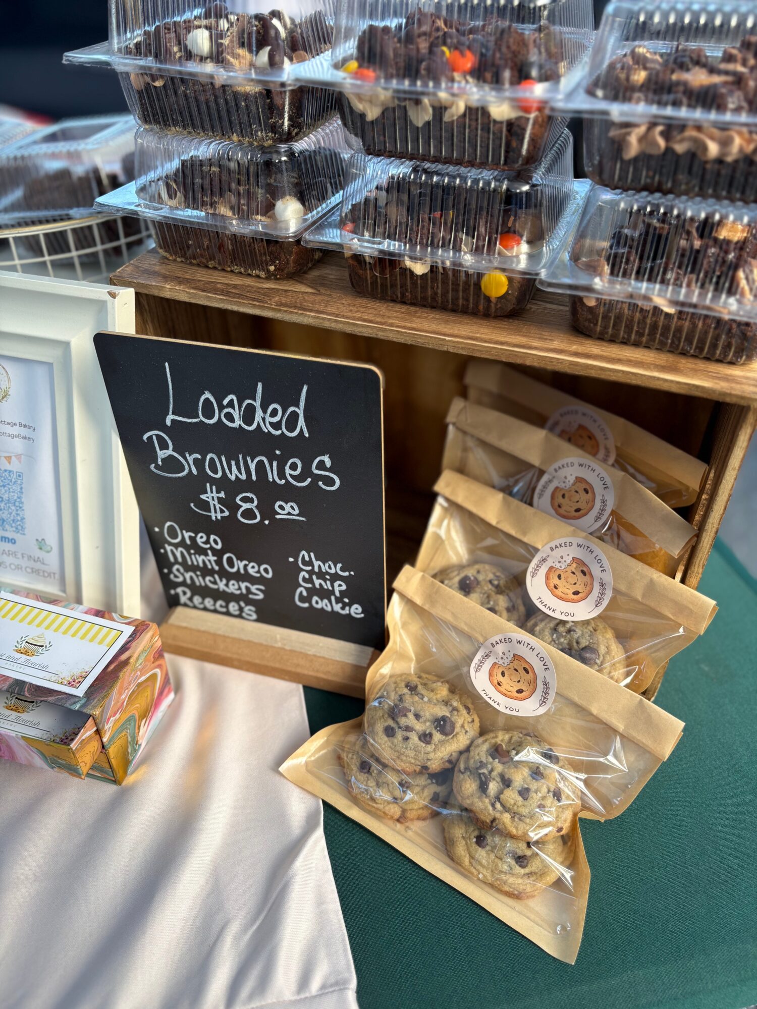 Display of loaded brownies in clear plastic containers and packaged cookies on a table, with a chalkboard sign listing flavors.