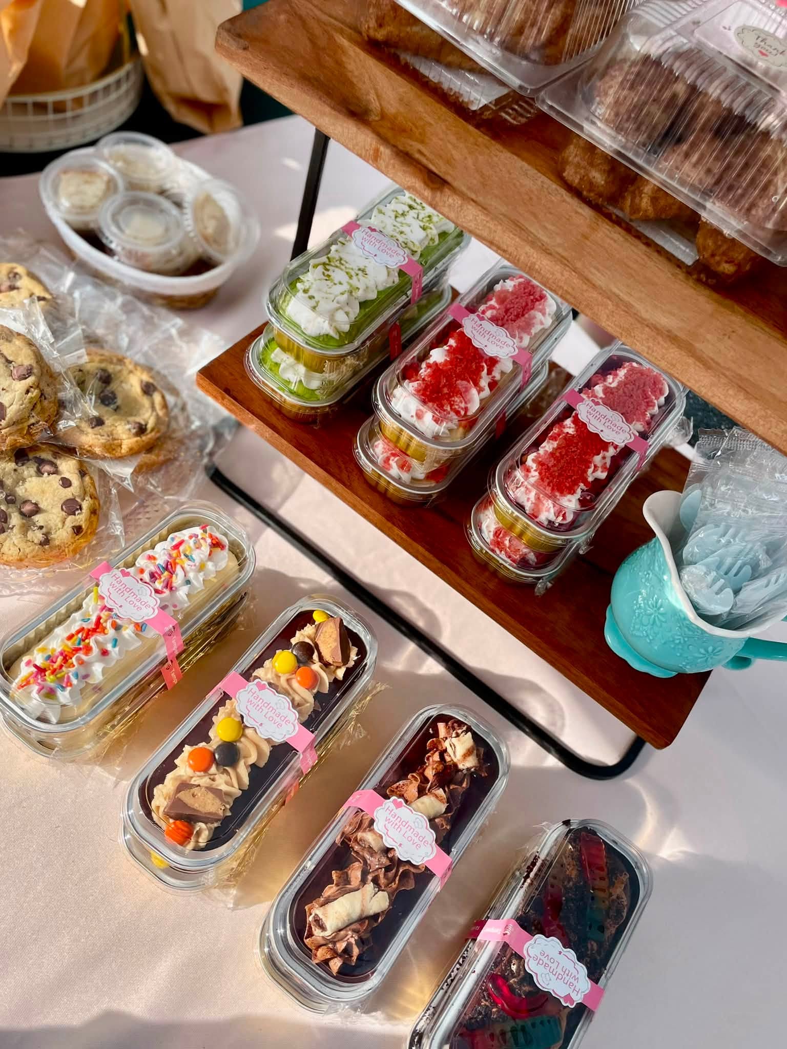 Assorted decorated cakes and desserts on a display table, with some in glass containers and others on a wooden stand.