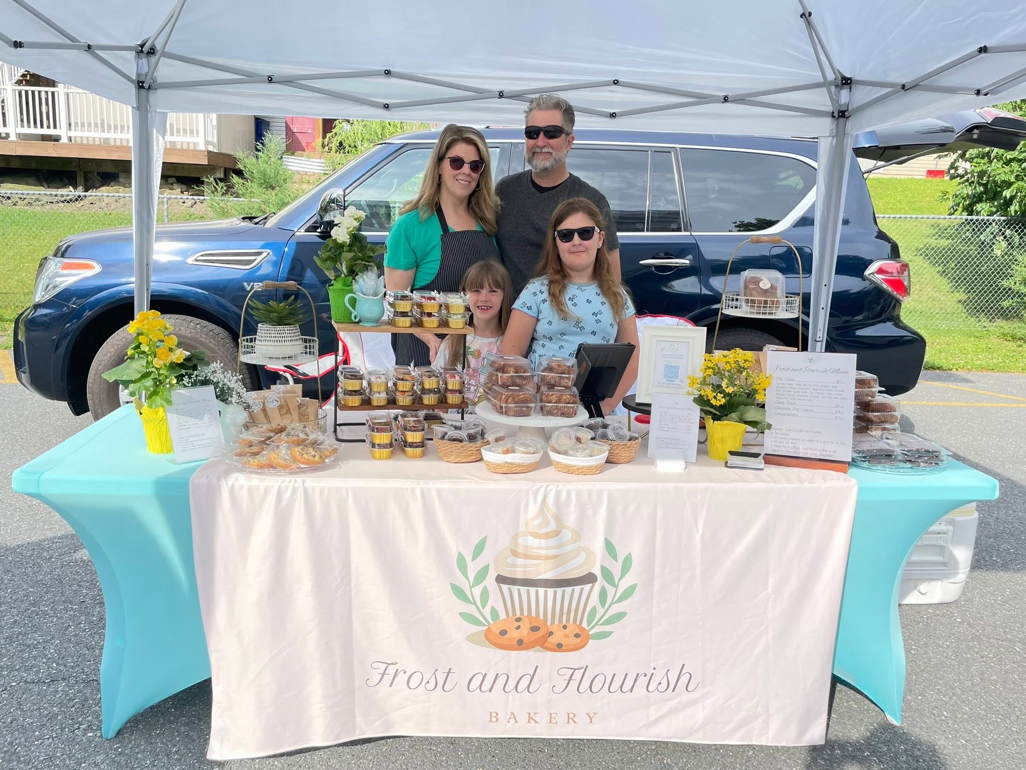 Family of four standing behind a table with baked goods at an outdoor event, under a white canopy, with a blue vehicle in background.
