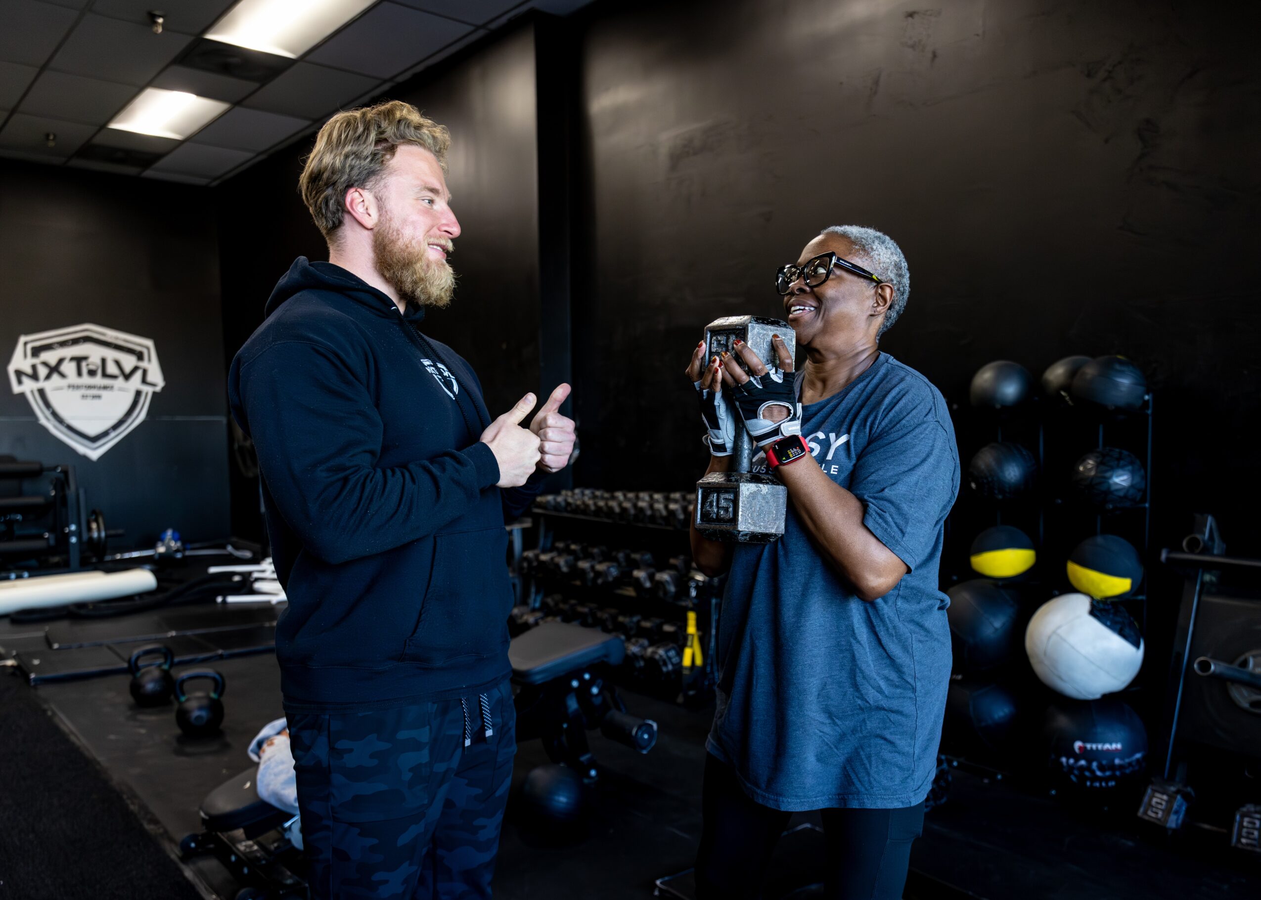 Two people talking in a gym, one holding a water bottle, with exercise equipment in the background.