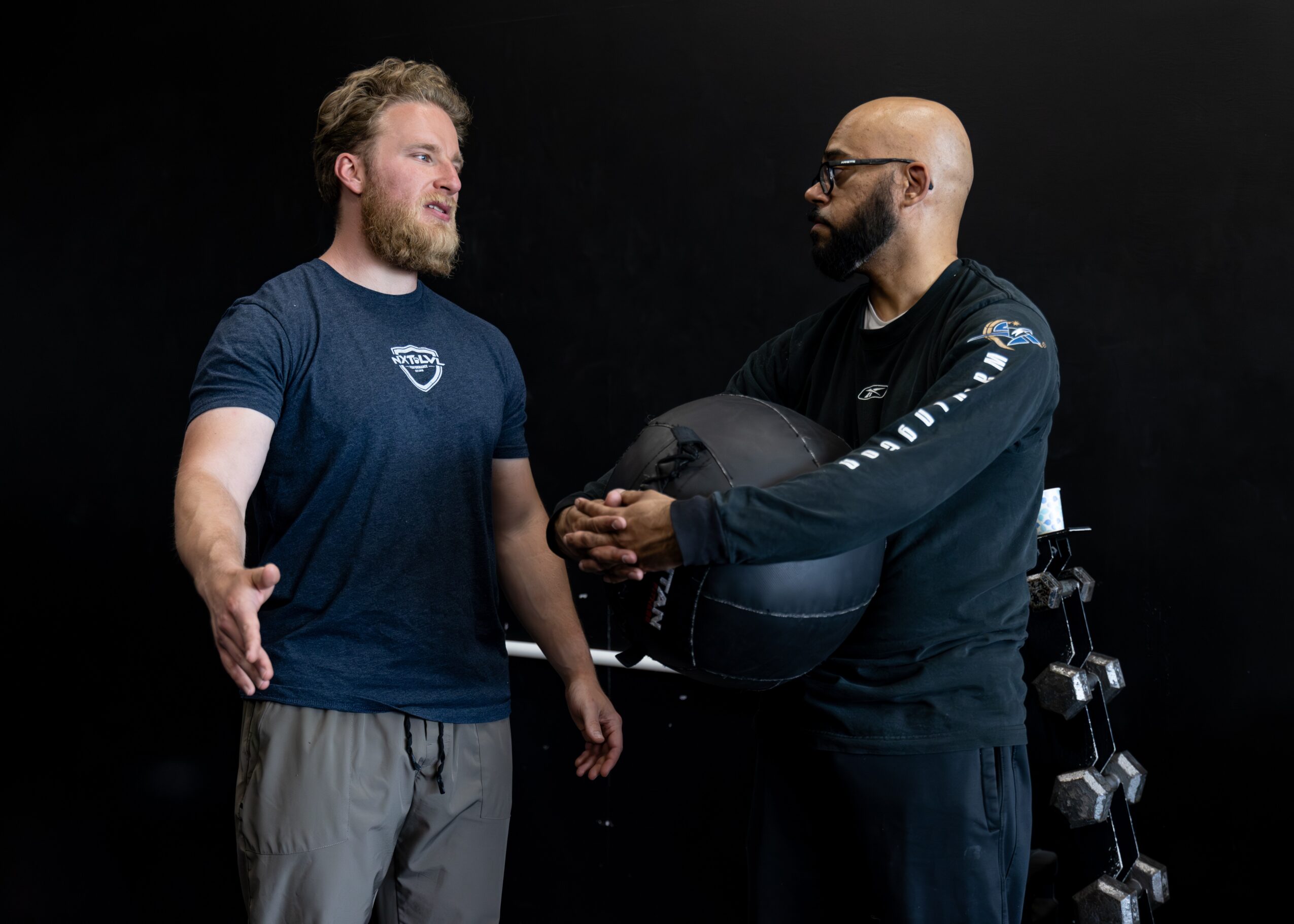 Two men standing and talking, one with a beard and wearing a blue shirt, the other with glasses and a black jacket, holding a helmet.