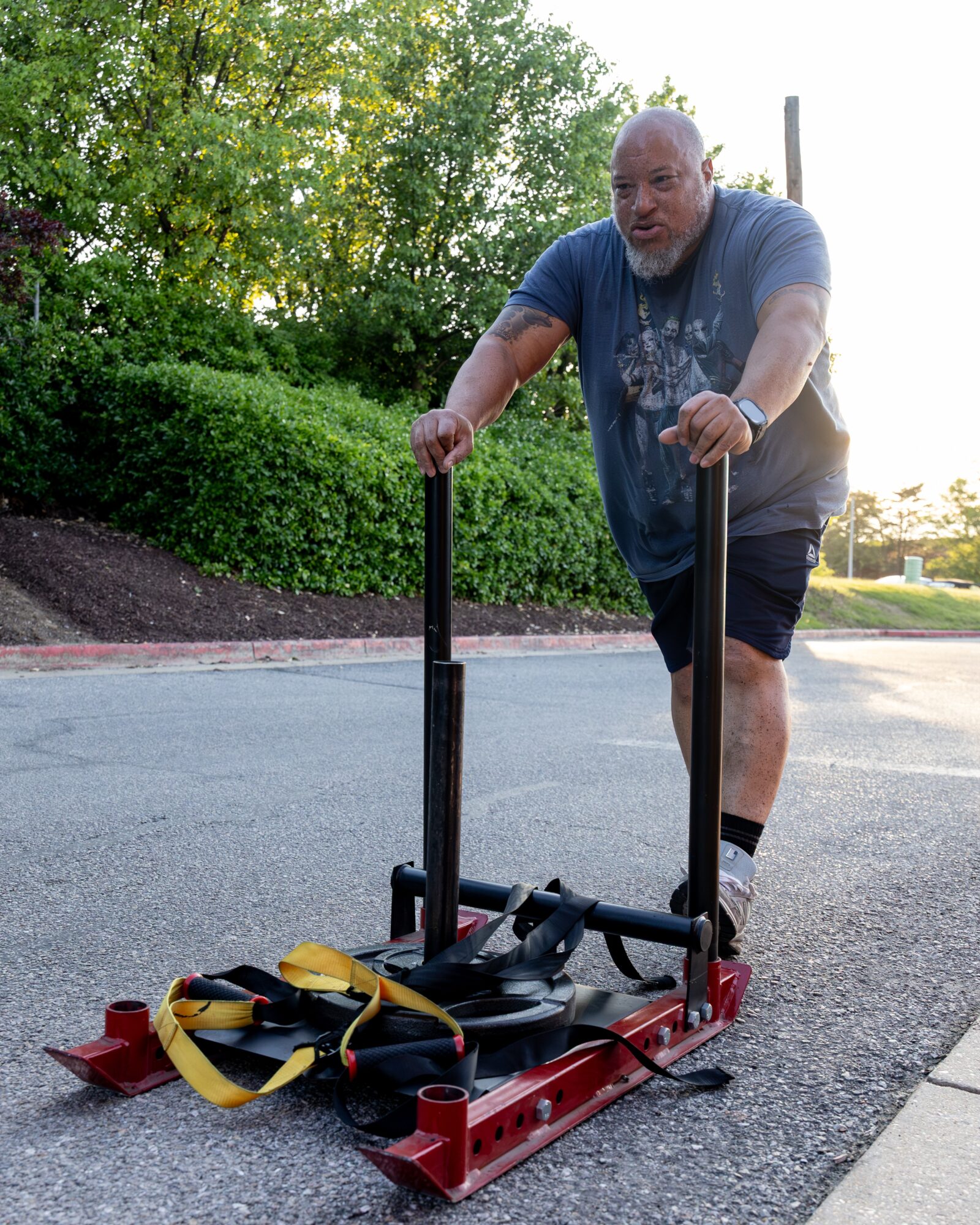 Man pushing a sled with exercise equipment on a paved outdoor path, surrounded by greenery and trees.
