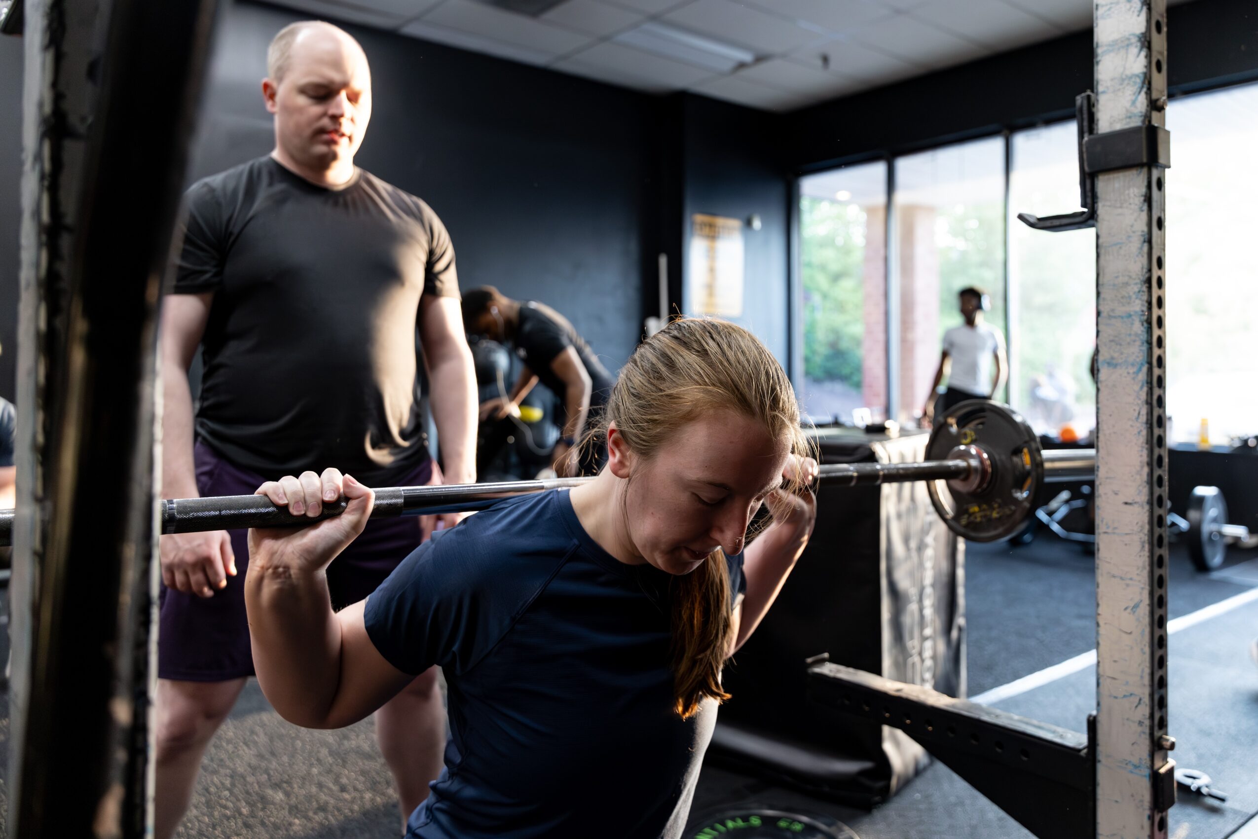 A woman lifts a barbell in a gym, with a man standing nearby and other people working out in the background.