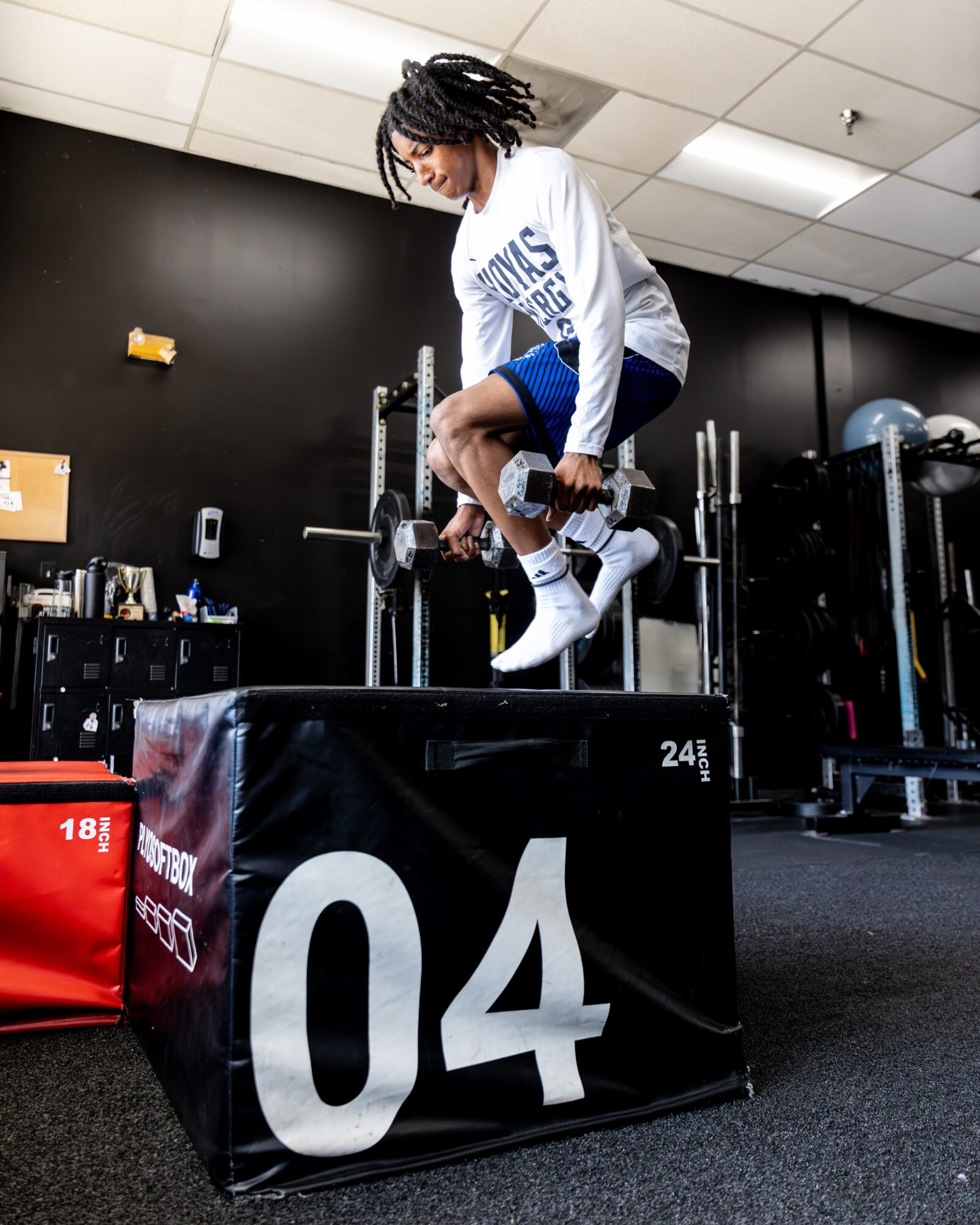 Person jumping onto a black box in a gym, wearing a white sweatshirt and blue shorts, with gym equipment in background.
