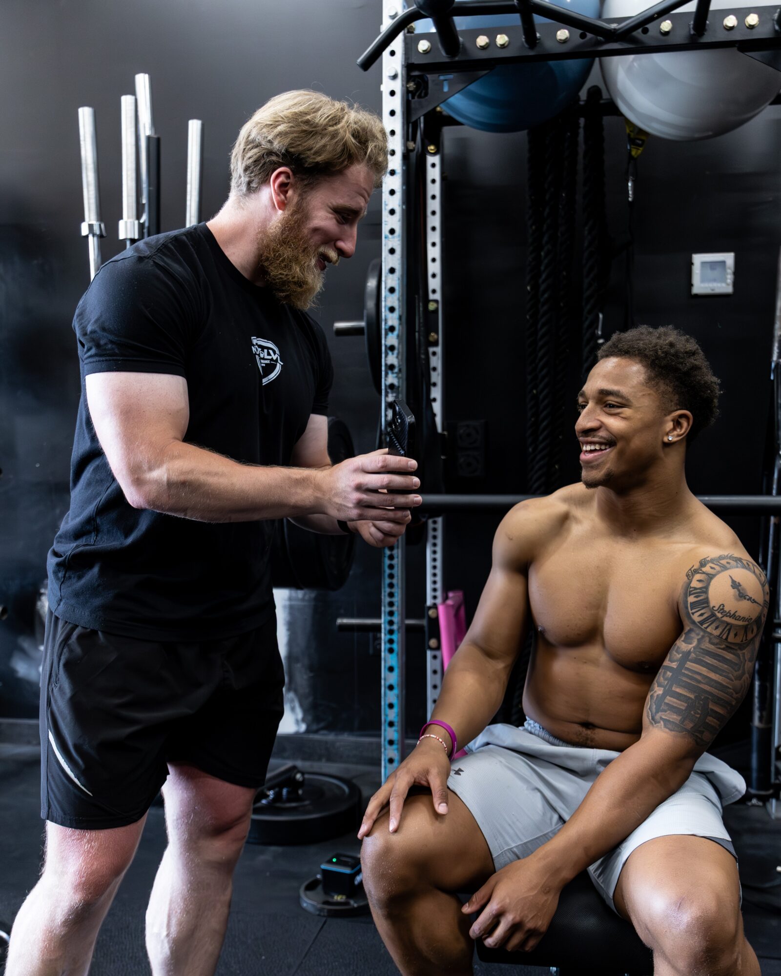 Two men in a gym, one shirtless with tattoos, smiling, the other in a black shirt, holding a barbell. Gym equipment visible.