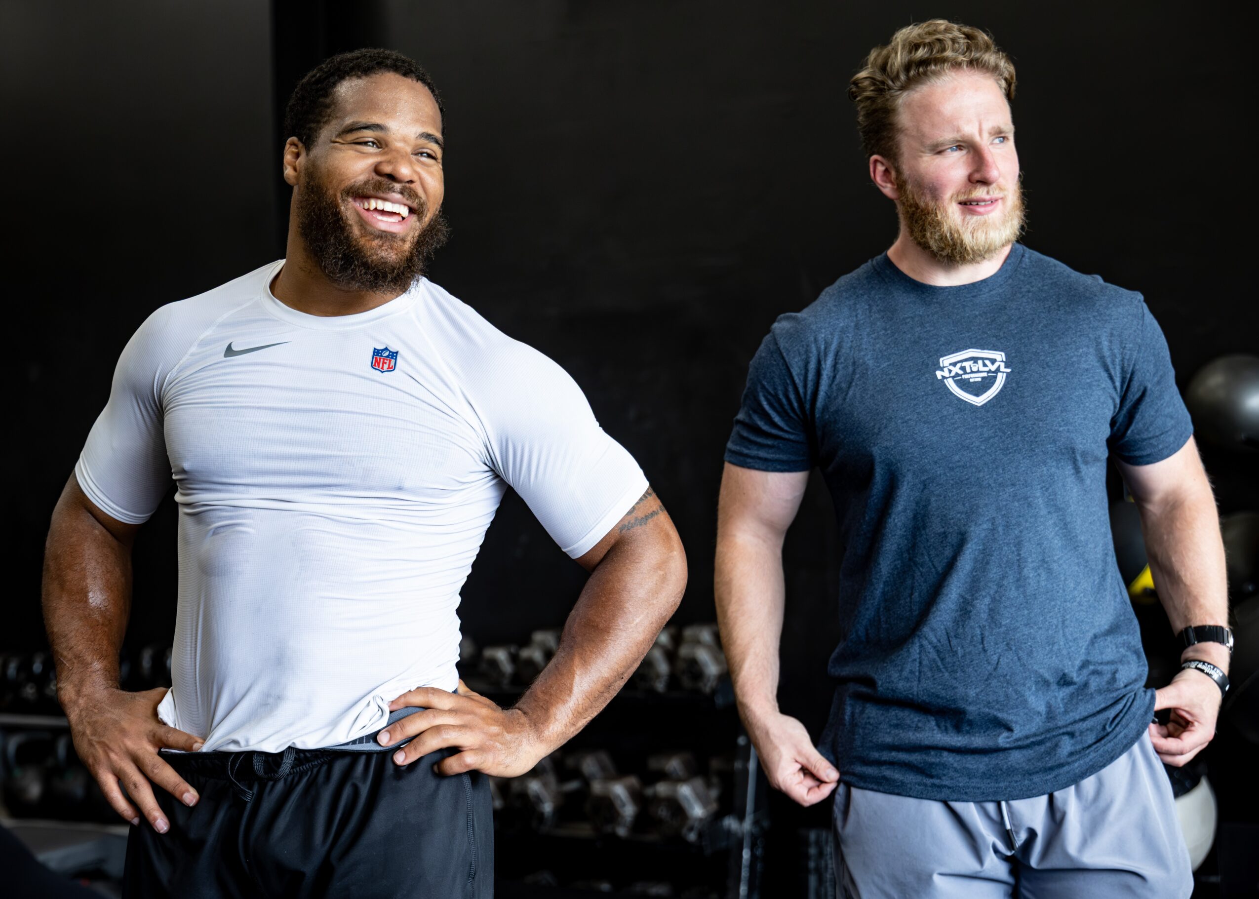 Two men standing side by side, smiling, in a dark indoor setting, one in a white sports shirt, the other in a blue t-shirt.