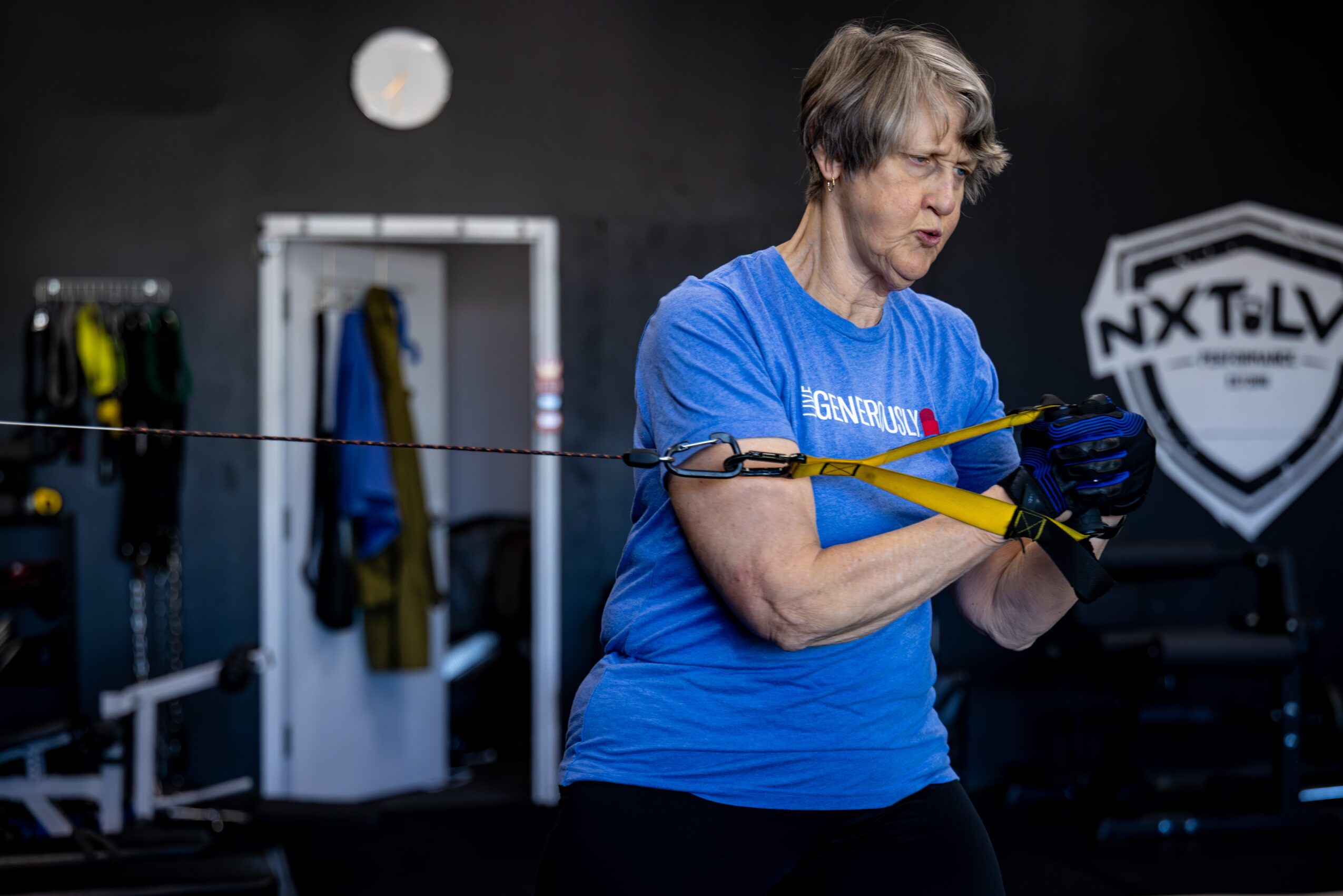 Older woman in blue shirt using resistance band in gym, with gym equipment and a logo in background.
