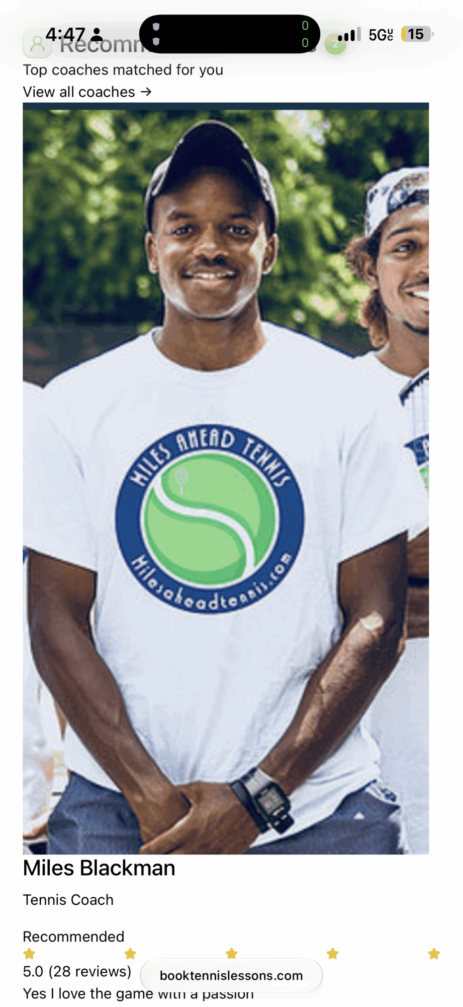 Man smiling outdoors wearing a white t-shirt with a green and blue logo, with trees in the background.