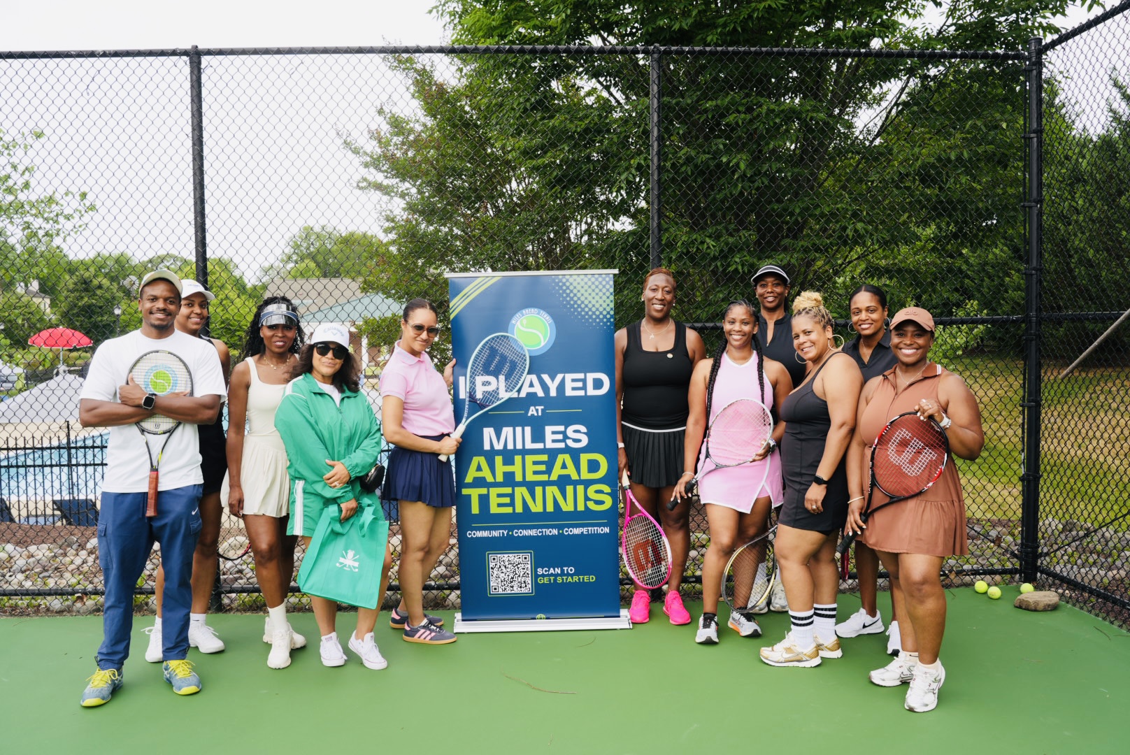 Group of people standing on a tennis court with a sign, trees, and a fence in the background.