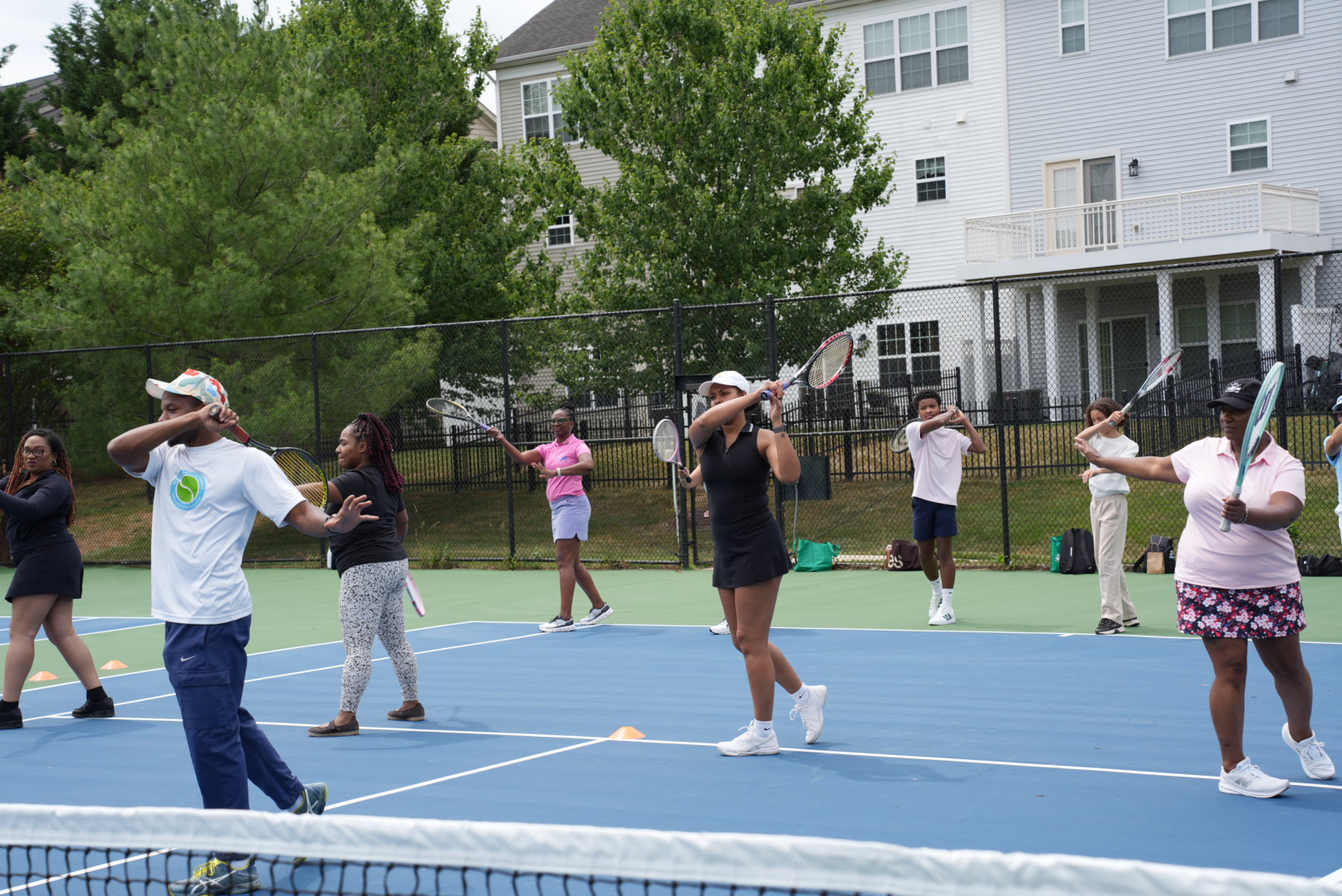 Group of children and adults practicing tennis on an outdoor court with trees and buildings in background.