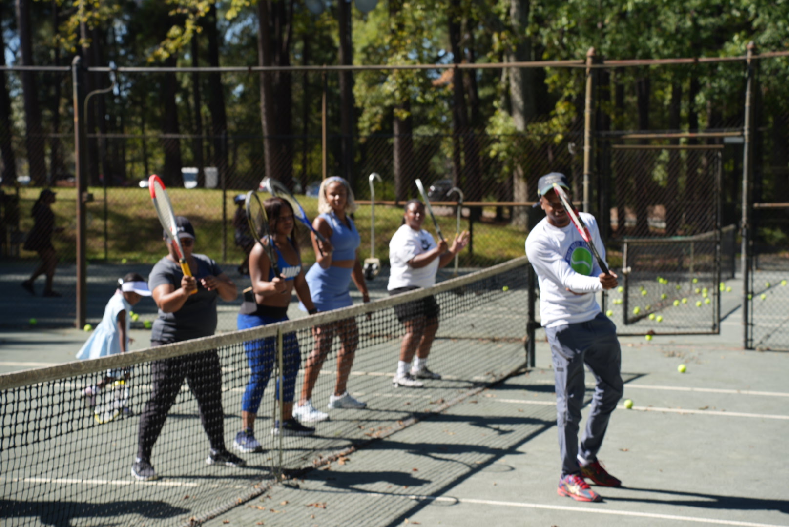 Group of people playing tennis on an outdoor court with trees in the background.