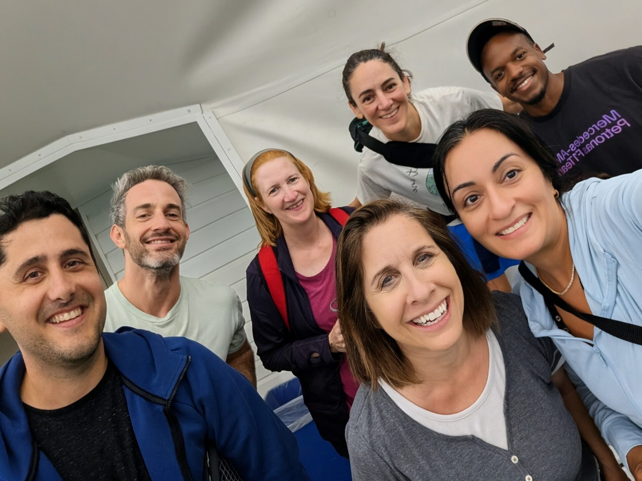 Group of seven smiling people taking a selfie indoors, some leaning in, with a white ceiling and wall in the background.