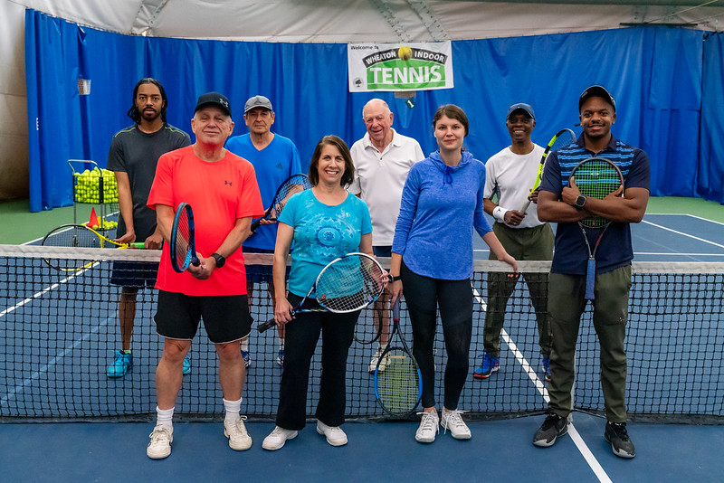 Group of eight people standing on a tennis court with a blue curtain background, holding tennis rackets, smiling, with a tennis sign above.