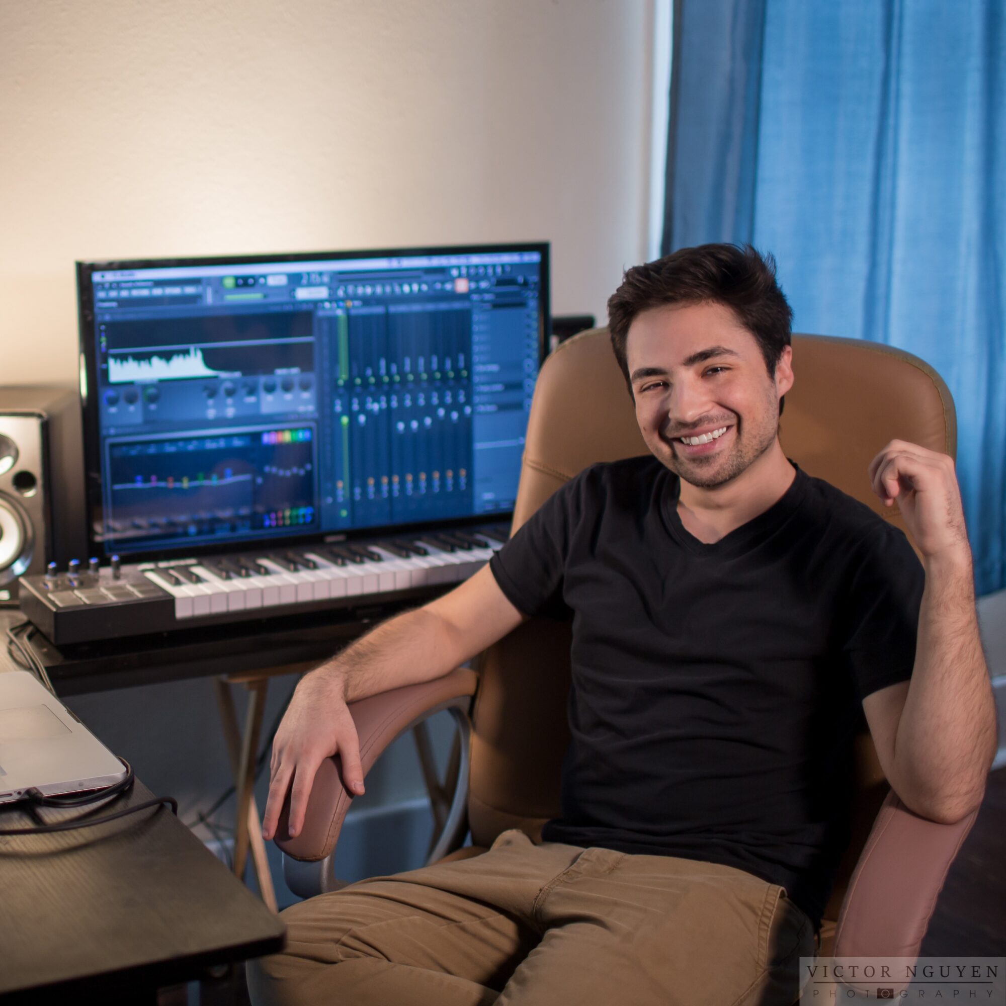 Young man sitting in a chair in front of a computer with music production software, smiling, in a room with a blue curtain.