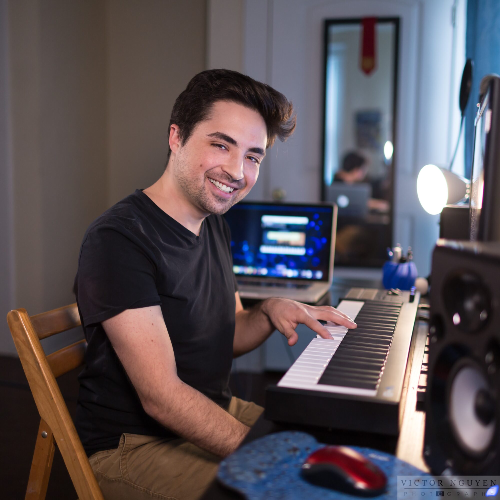 Young man smiling at keyboard in a room with a computer monitor and mirror, sitting on a wooden chair.