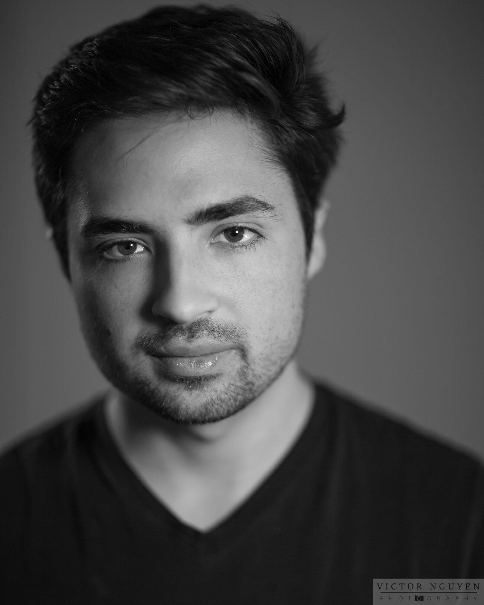 Black and white portrait of a young man with dark hair, slight beard, wearing a V-neck shirt, looking at the camera.