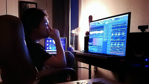 Person sitting at a desk, working on a computer with a music editing program, in a dimly lit room.