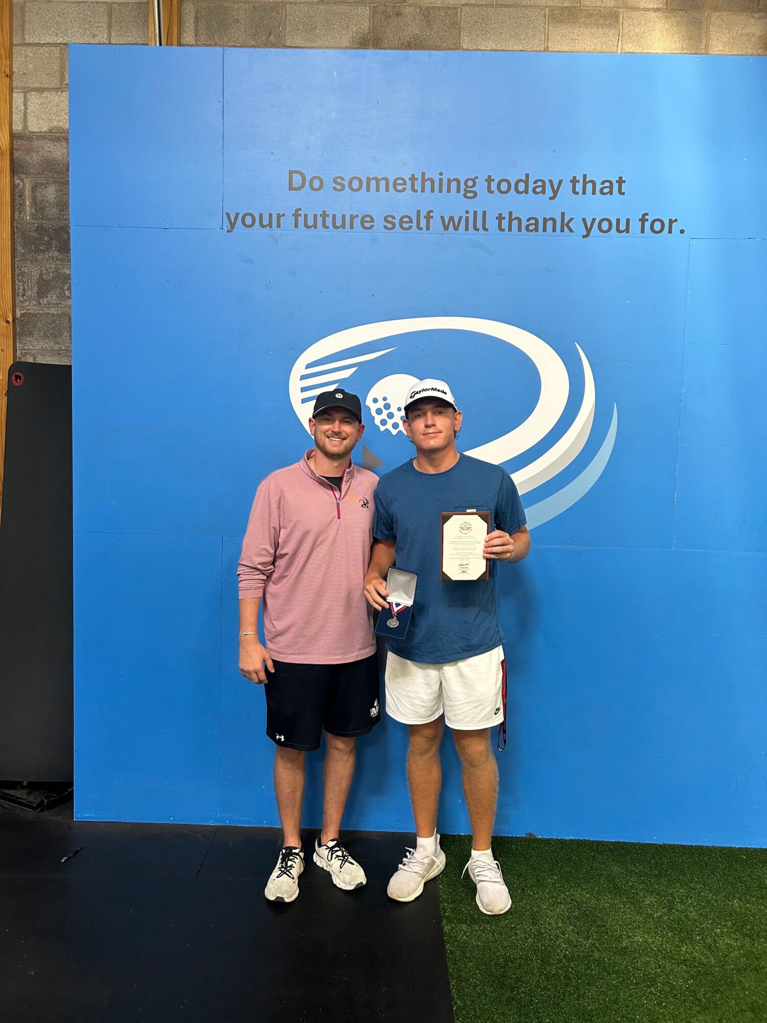 Two women standing in front of a blue backdrop with a logo and text, one holding a plaque, both smiling, outdoors.