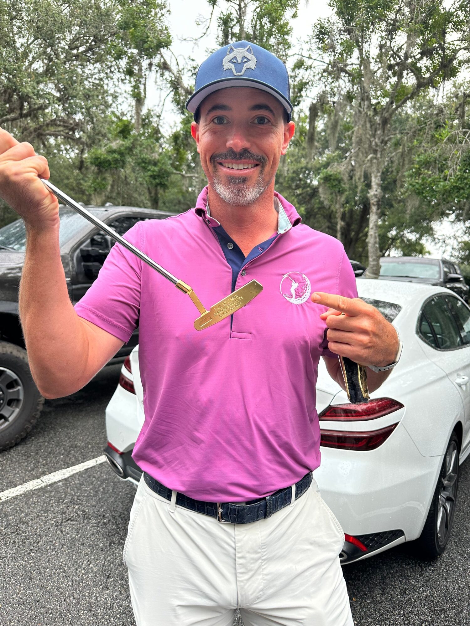 Man in pink shirt holding golf club, smiling, standing next to a white car outdoors with trees in background.