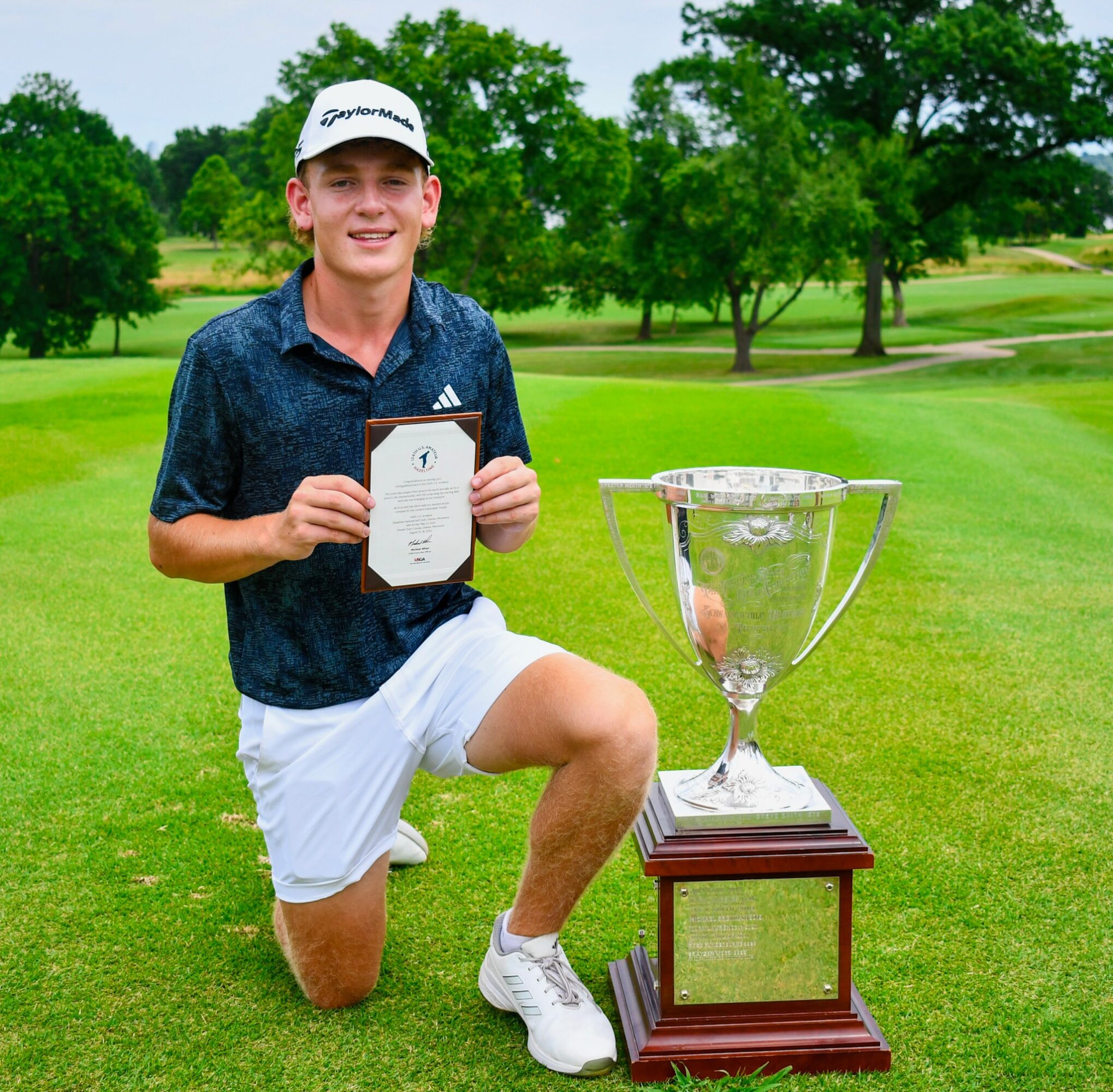 Young man kneeling on grass holding a certificate next to a large trophy on a golf course with trees in the background.