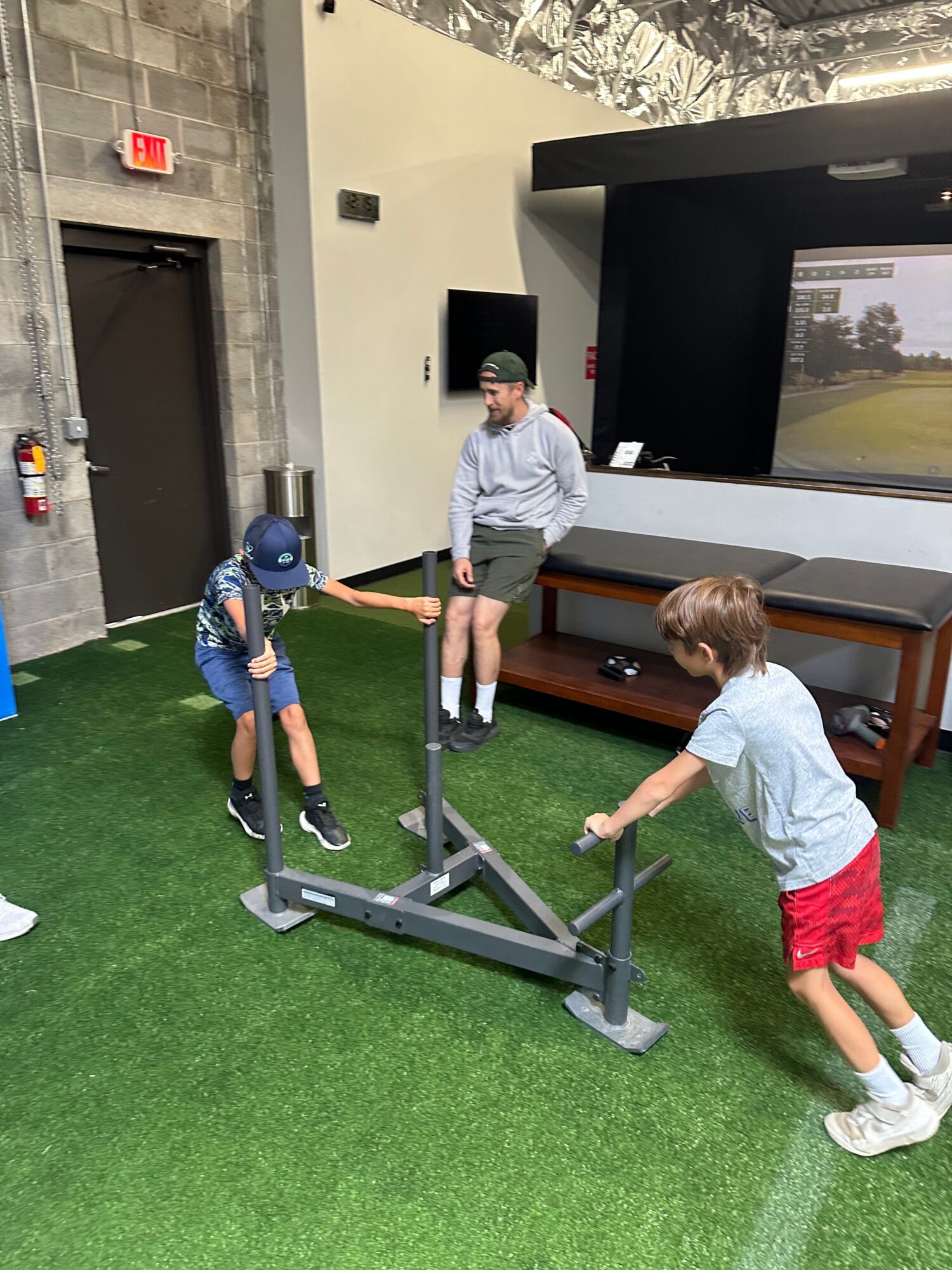 Two children playing with a treadmill indoors, one standing and the other sitting on the floor, on green carpet.