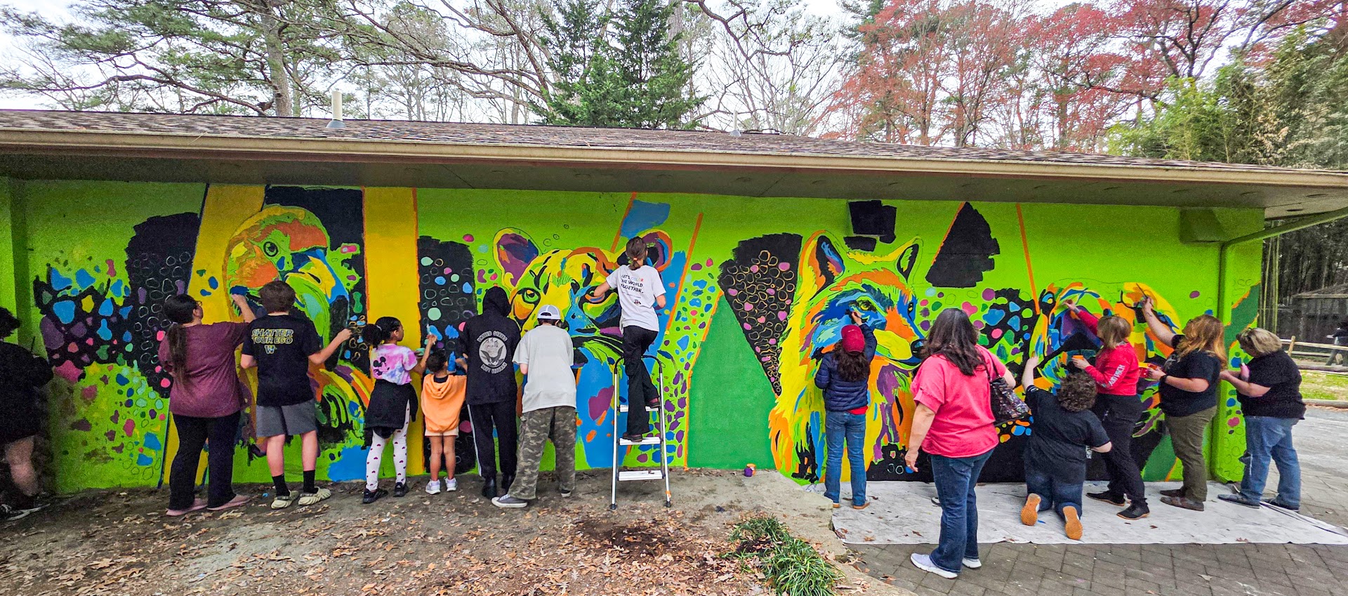 Group of people painting a colorful mural on a wall outdoors, with trees in the background.