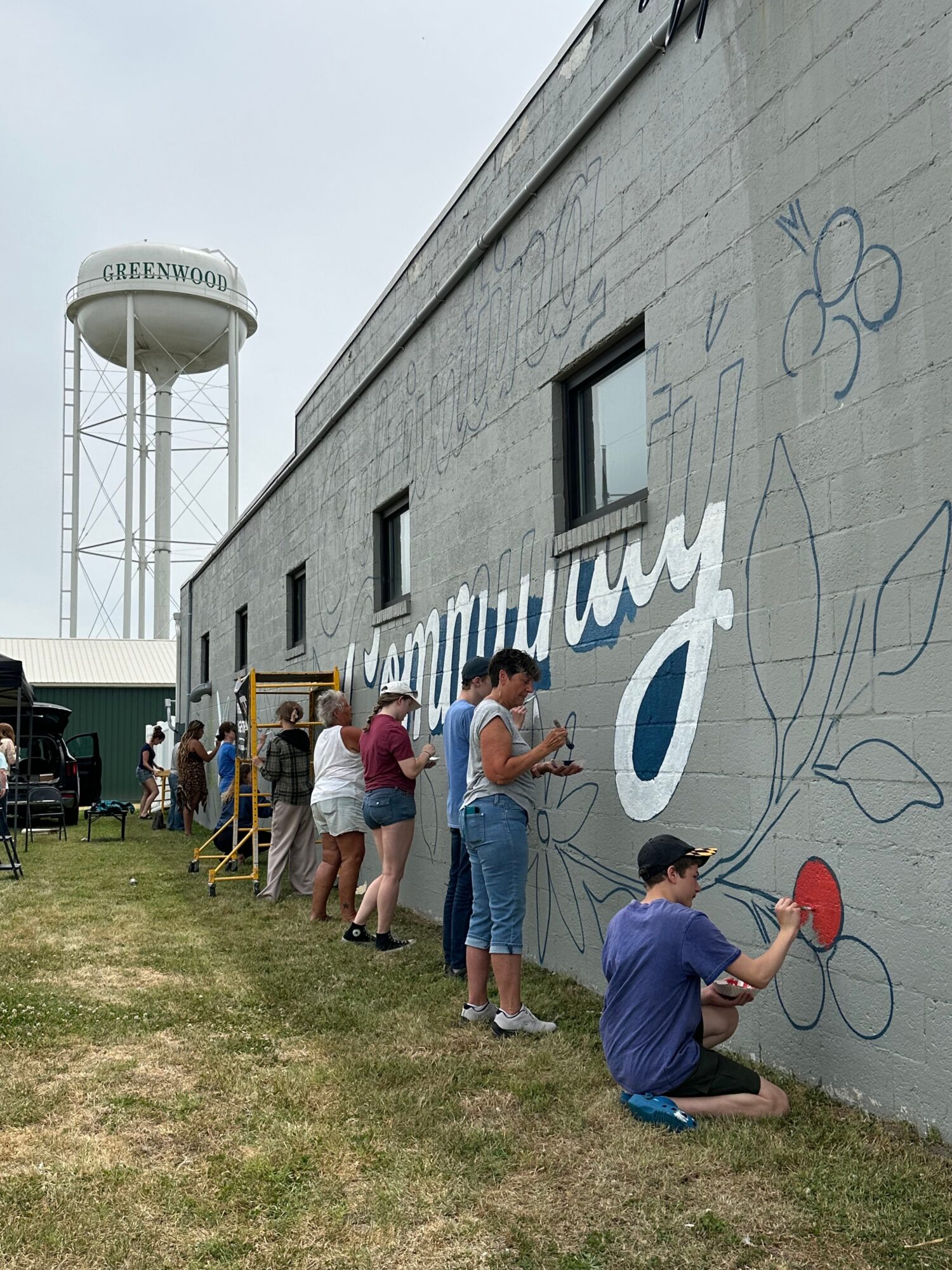 People painting a mural on a gray building wall with a water tower labeled 'GREENWOOD' in the background.