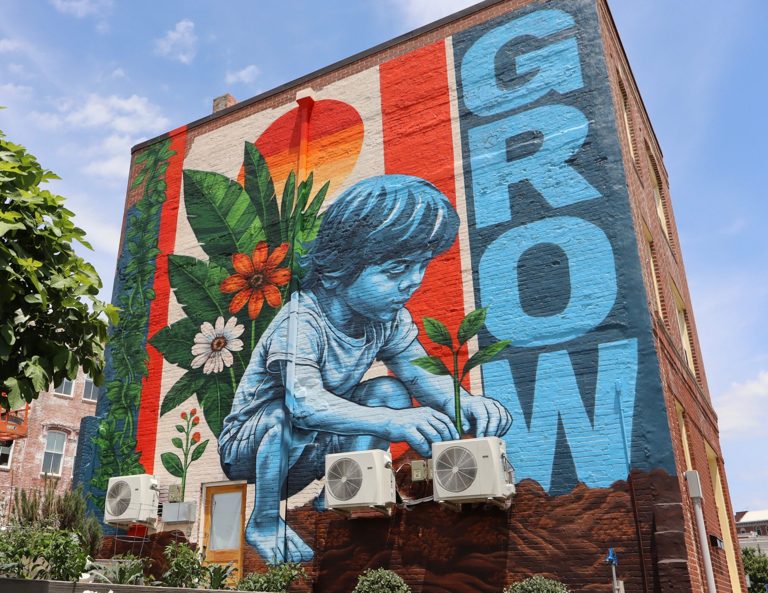 Large mural of a girl planting a seed, with colorful flowers and the word GROW on a brick building wall.