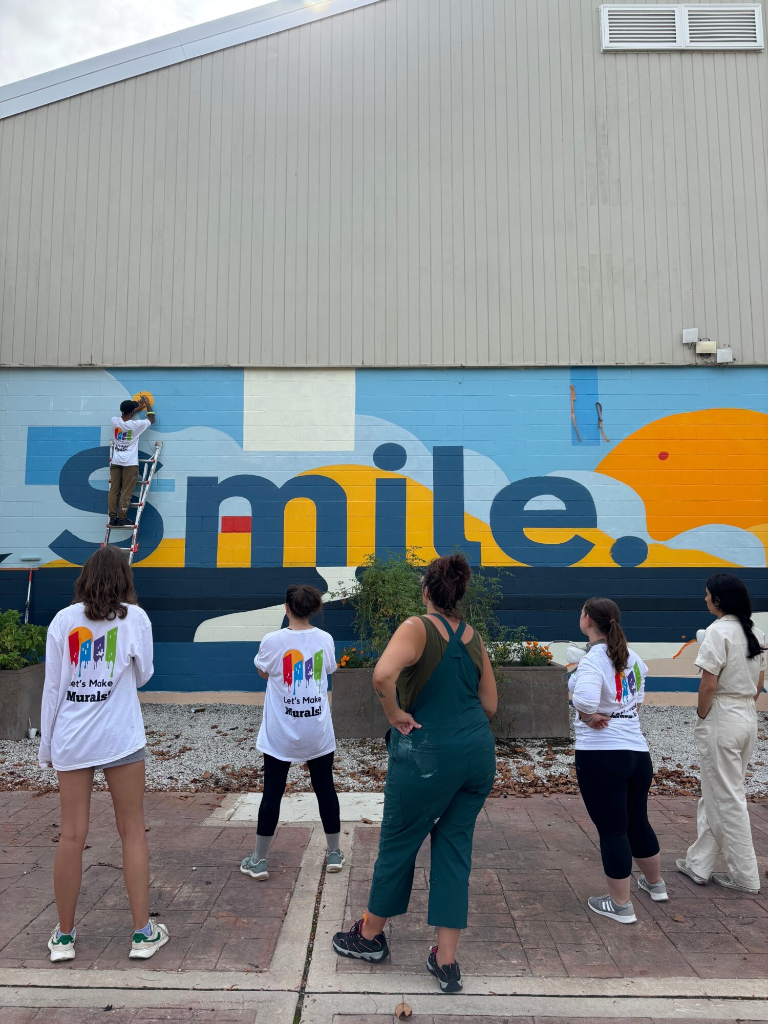 Group of people standing in front of a colorful mural with the word 'smile' and a yellow shape, outdoors.