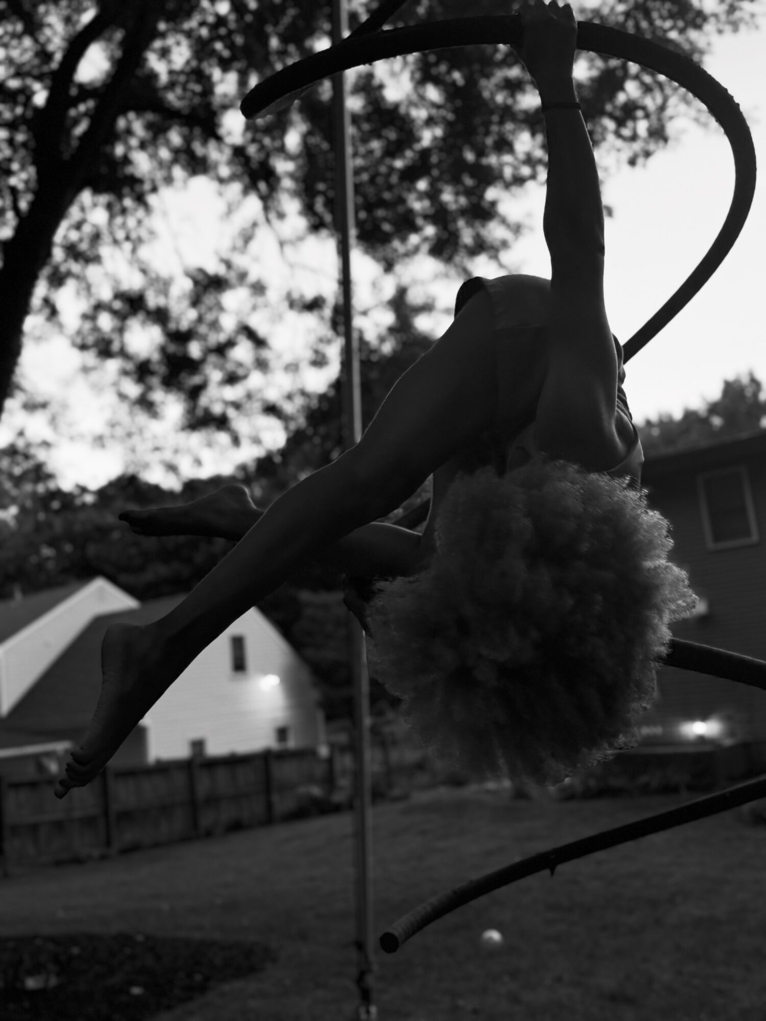 Person with curly hair hanging upside down on a hoop outdoors, trees and houses in background, black and white.