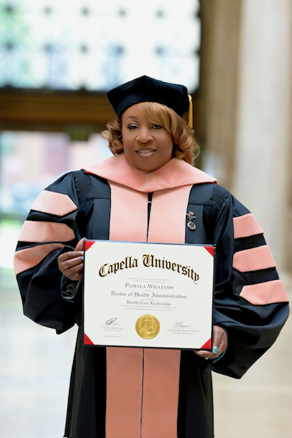 Woman in graduation gown and cap holding diploma, smiling, indoors with blurred background.