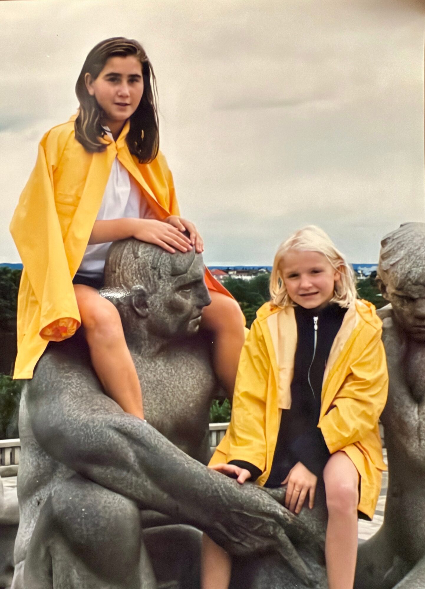 Two girls with yellow jackets sit on stone sculptures outdoors, with a cloudy sky in the background.
