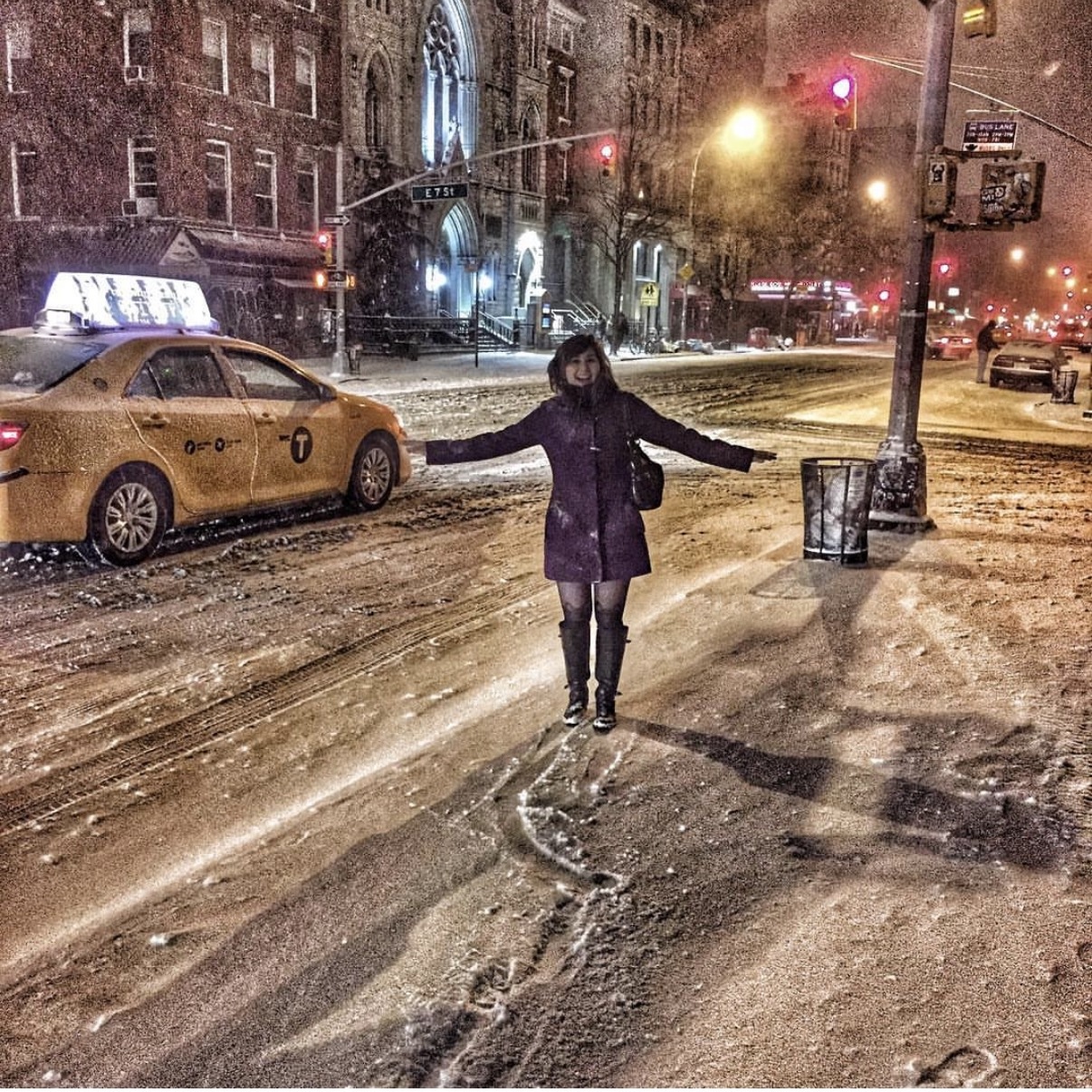 Person standing in snowy street at night with arms outstretched, city buildings and cars in background, streetlights illuminating scene.