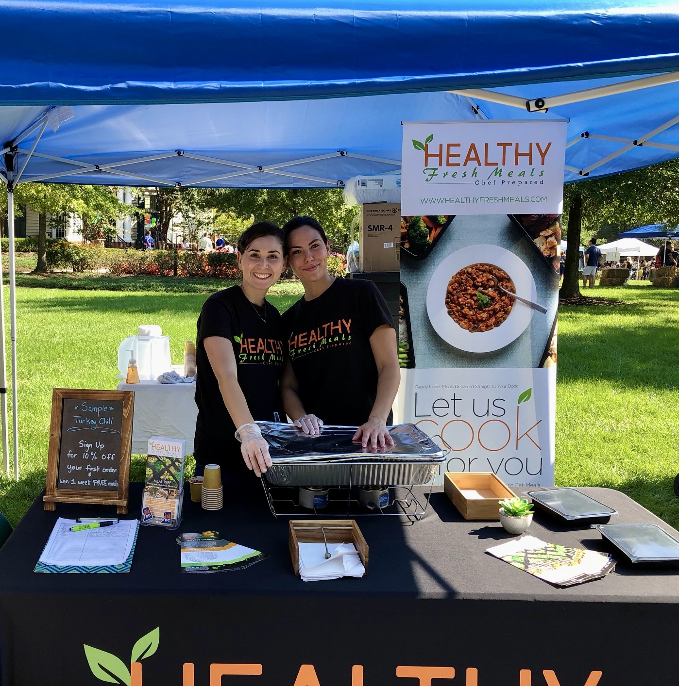 Two women stand behind a table at an outdoor health food booth, smiling, with a sign and food items displayed.