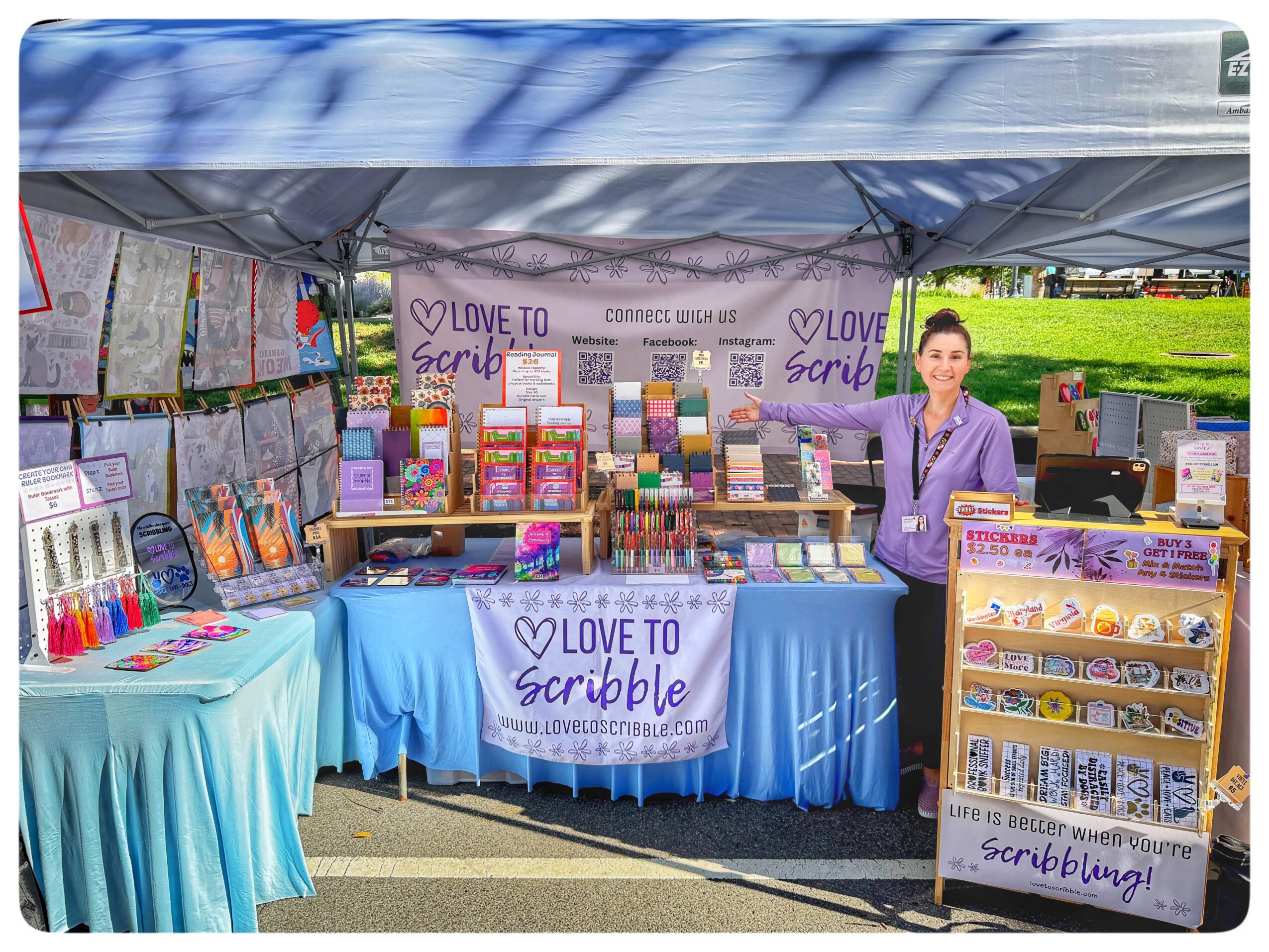 Outdoor booth selling colorful stationery and craft supplies, with a woman standing beside the display, under a canopy.