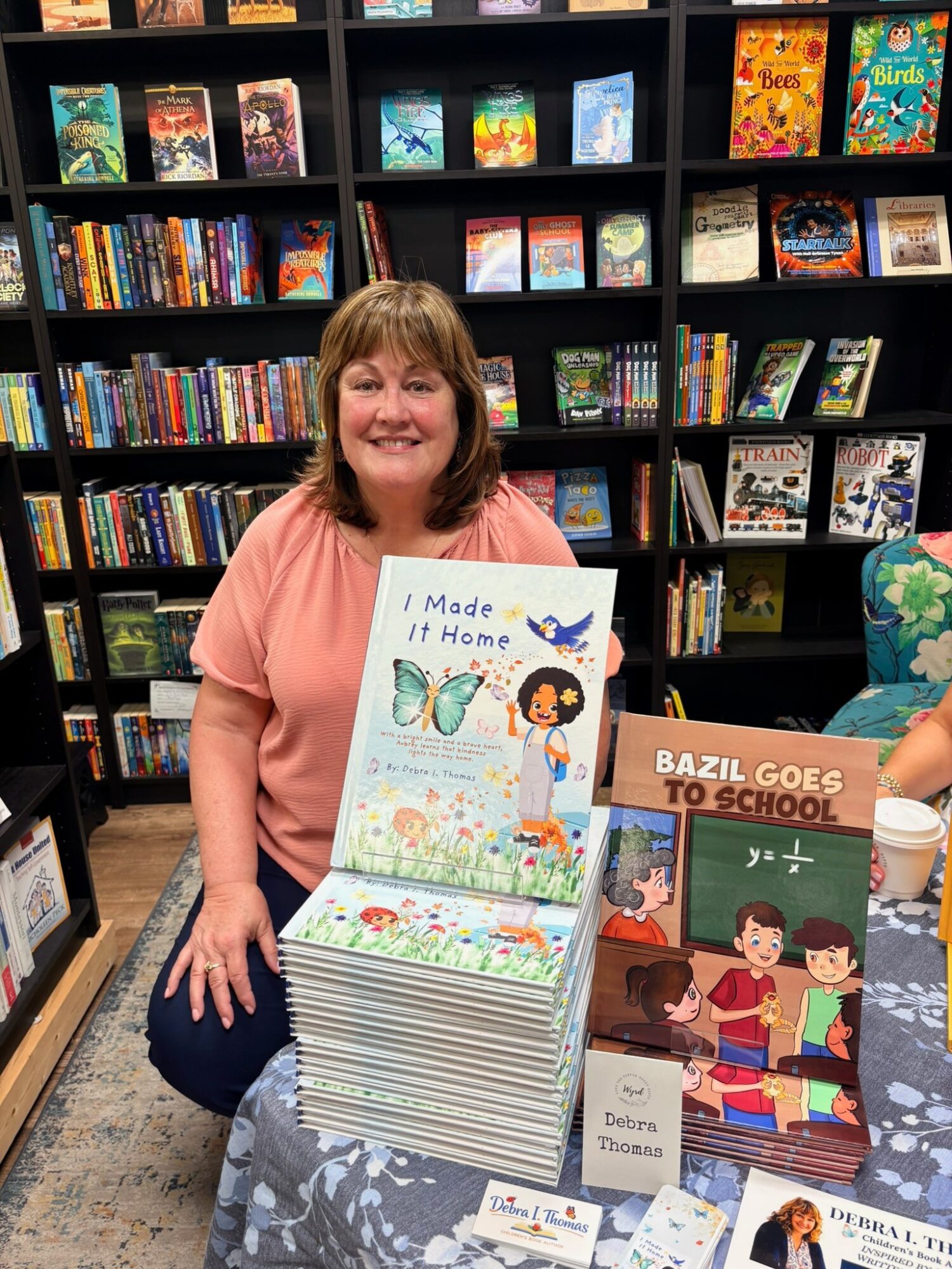 Woman sitting at a table with children's books in a bookstore, smiling, with bookshelves in the background.