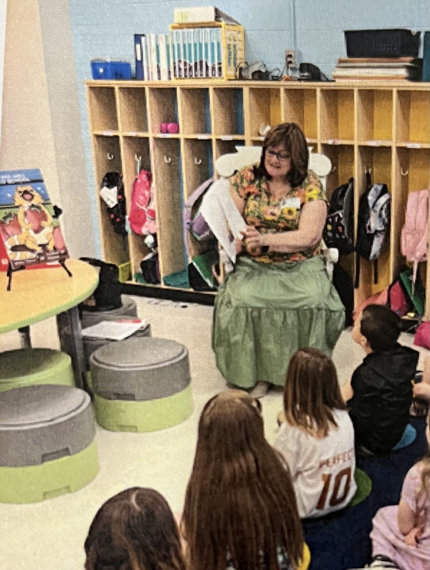 Woman sitting on a chair with children sitting on the floor in a classroom with cubbies and colorful items.