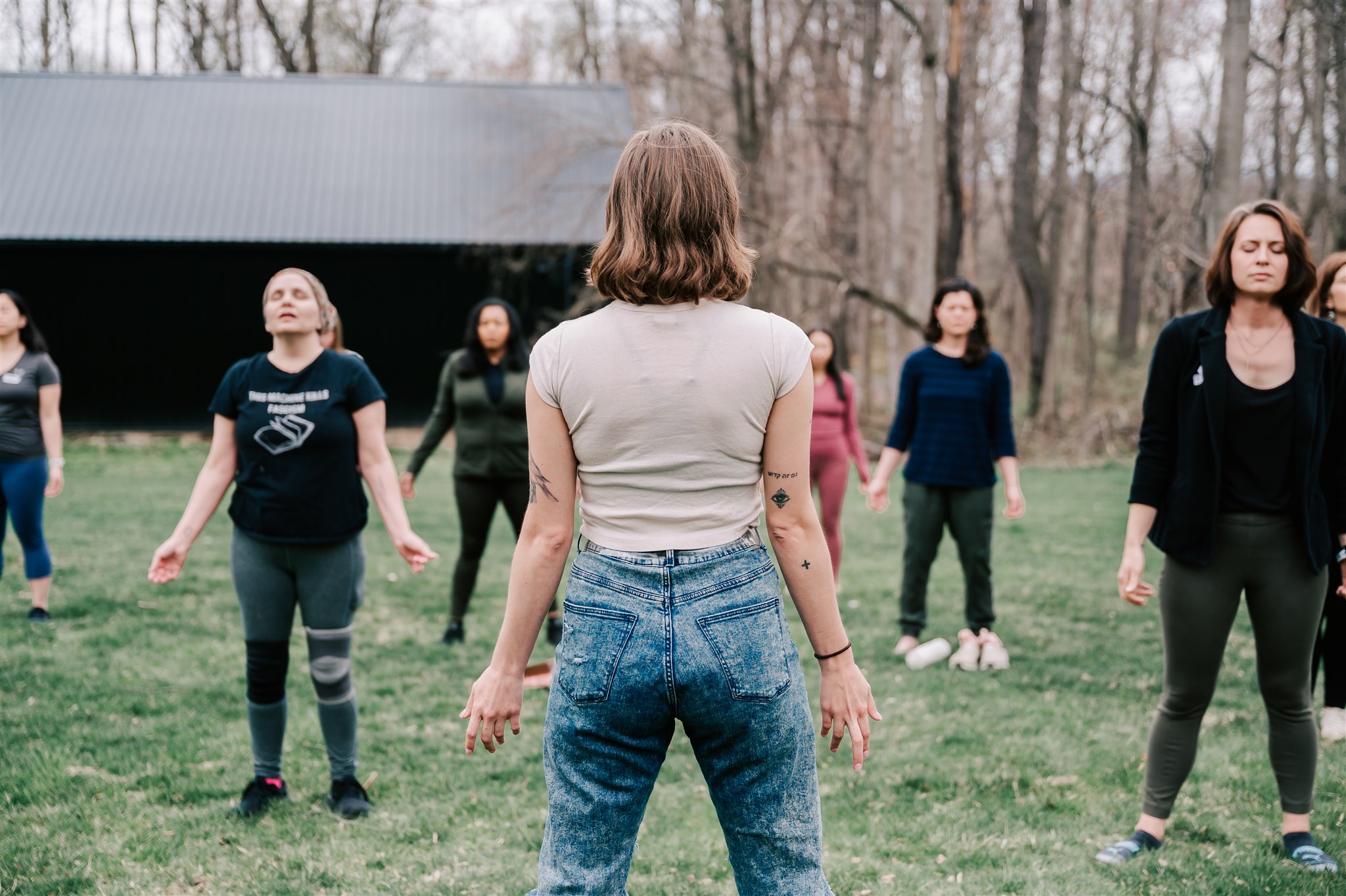 Group of women standing outdoors on grass, facing a woman with back to camera, trees in background.