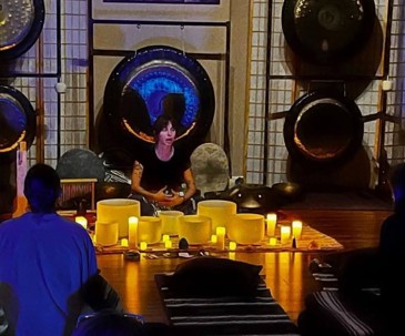 Person standing behind a table with yellow candles, with large gong and drums in background, audience seated nearby.