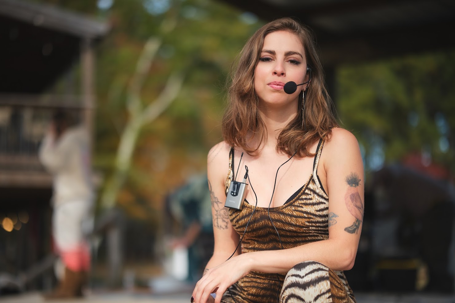 Young woman with shoulder-length hair wearing a tiger-striped top and a headset microphone, standing outdoors with trees in the background.