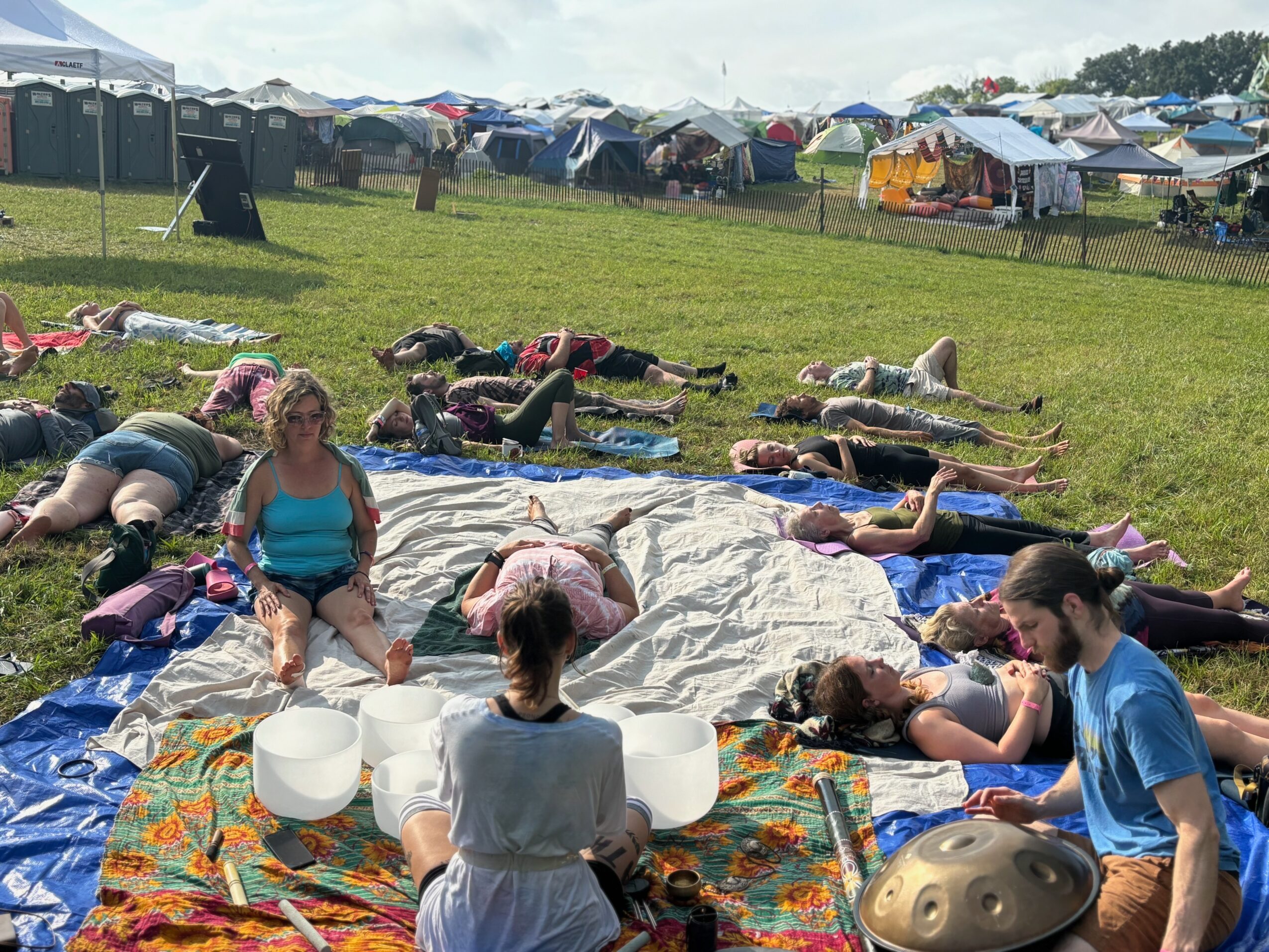 Group of people lying on mats and blankets outdoors, participating in a yoga or meditation session on a grassy field with tents in the background.