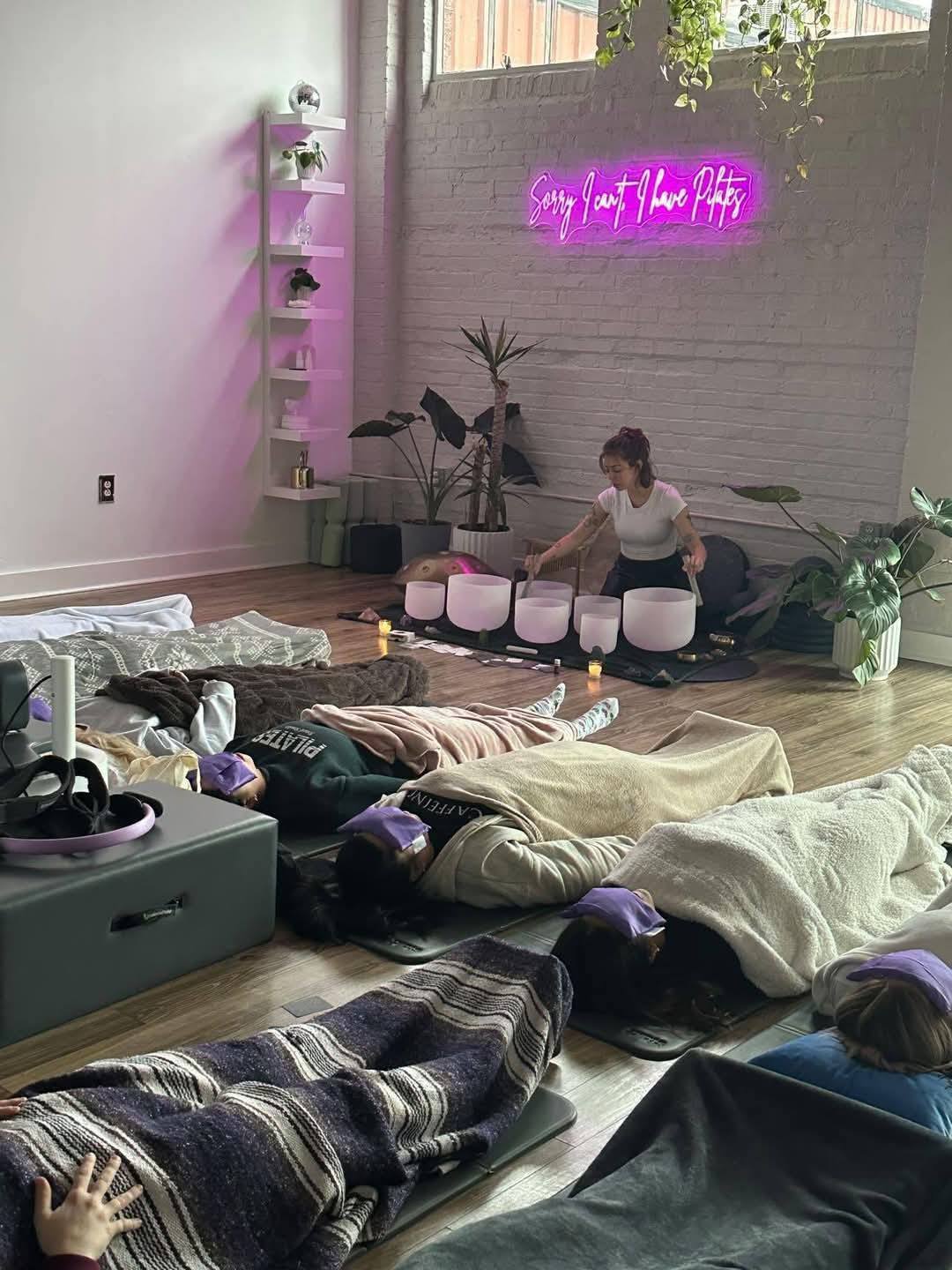 Person lying on the floor with others, neon sign on wall, plants, and shelves in room, with various objects scattered around.