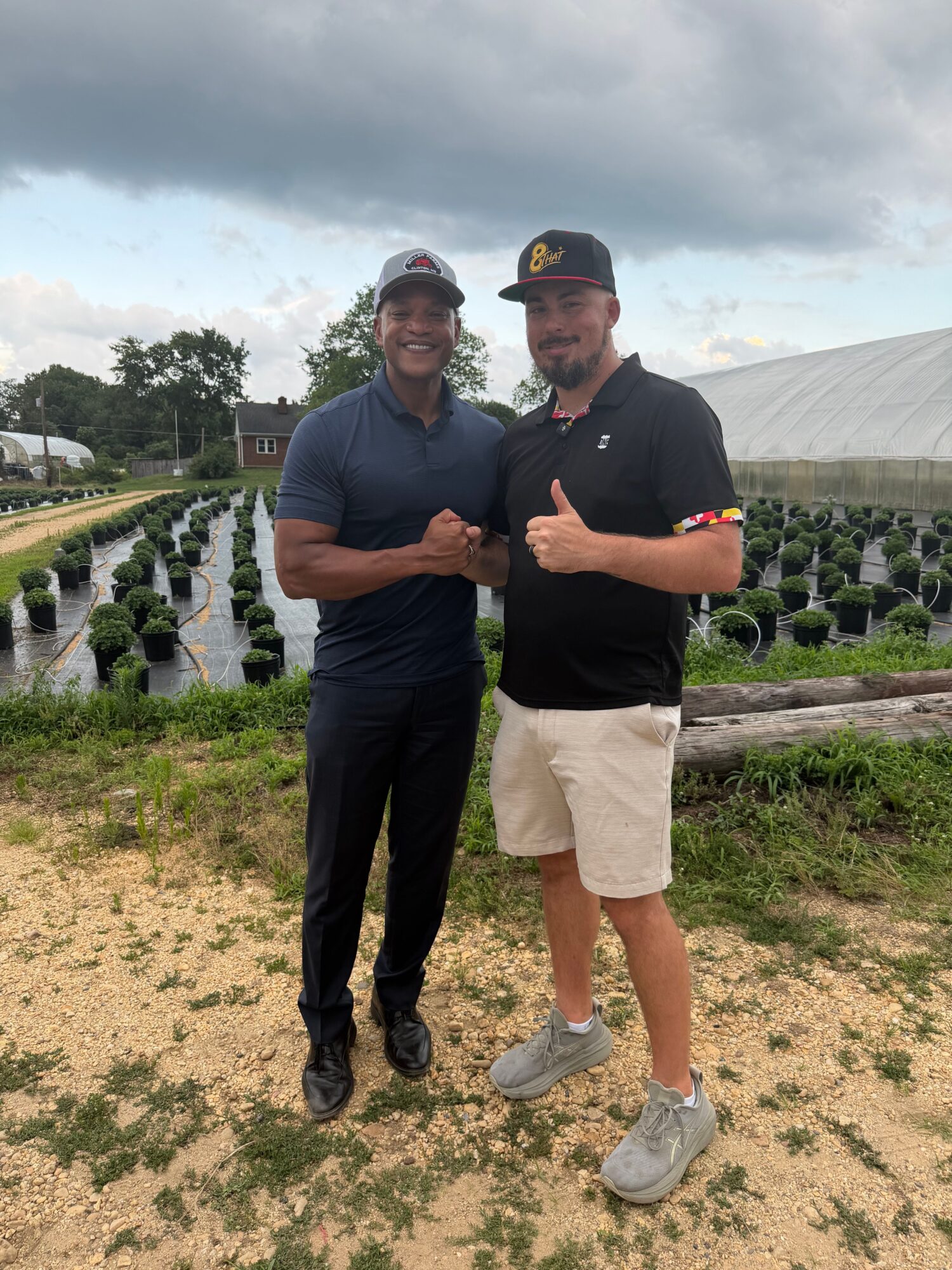 Two men standing outdoors on a grassy area, one with a black shirt and beige shorts, the other with a dark shirt and black pants, holding hands and smiling.