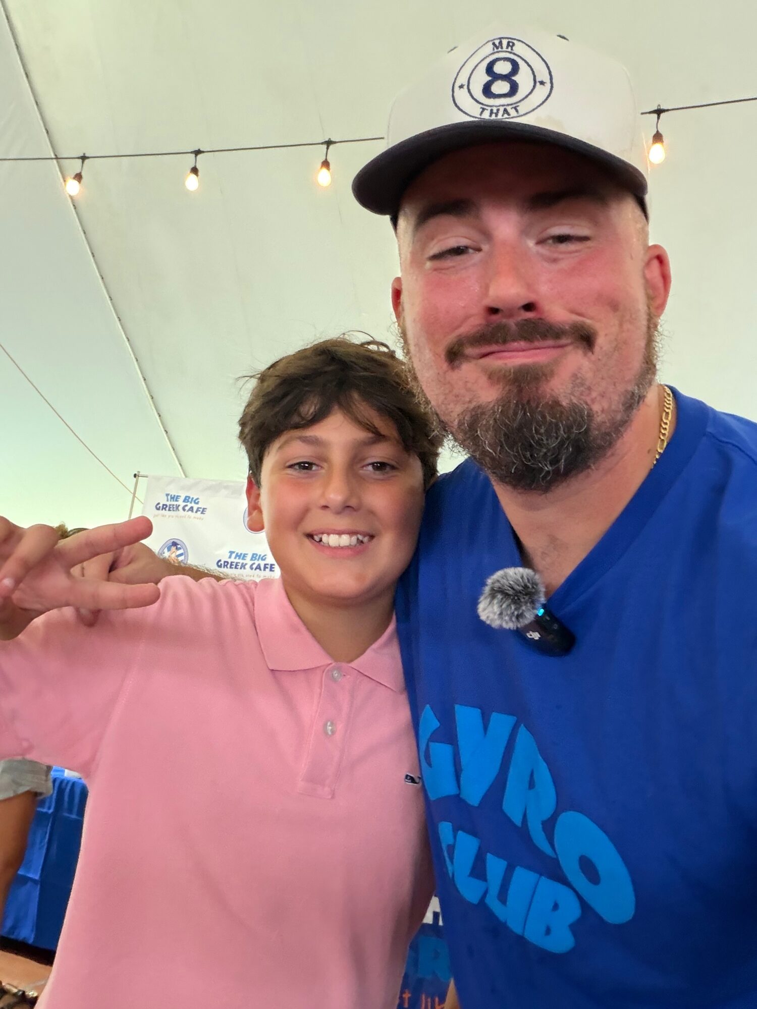 A man and a boy smiling, taking a selfie indoors with string lights overhead, man wearing a cap and blue shirt, boy in pink shirt.