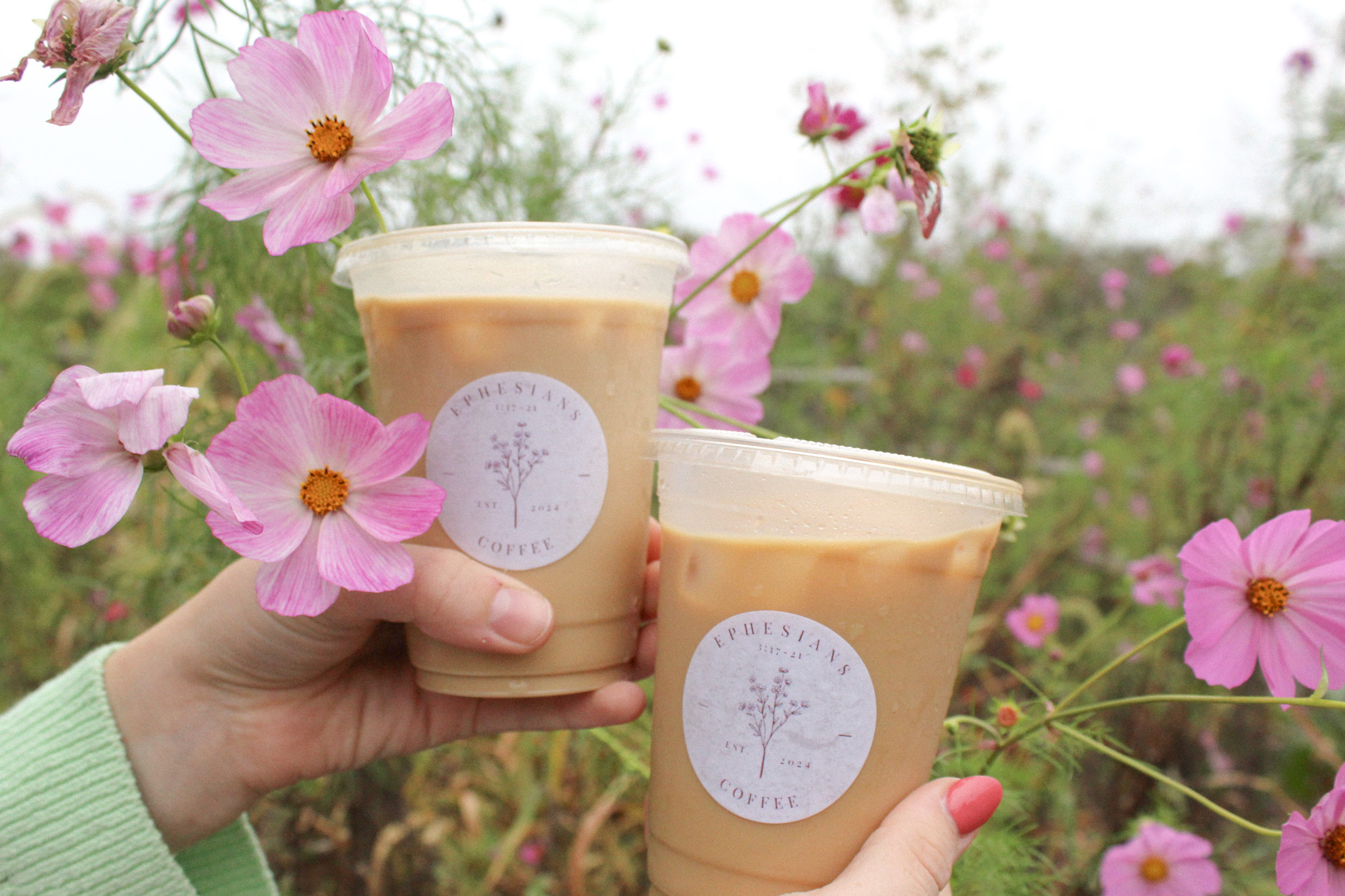 Person holding two cups of coffee or tea outdoors with pink flowers in background.