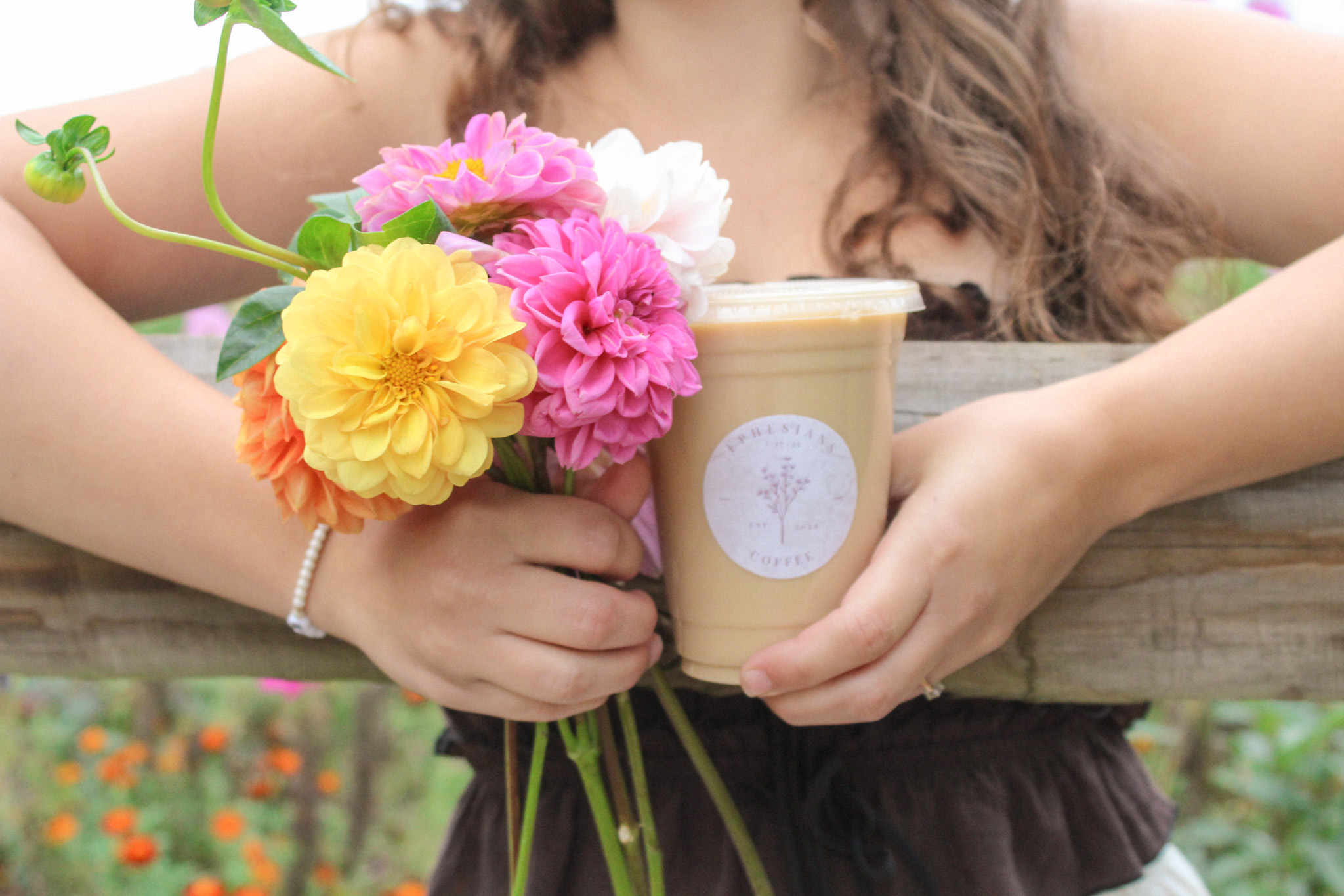 Person holding a cup with flowers and a small bouquet, outdoors with greenery and flowers in the background.