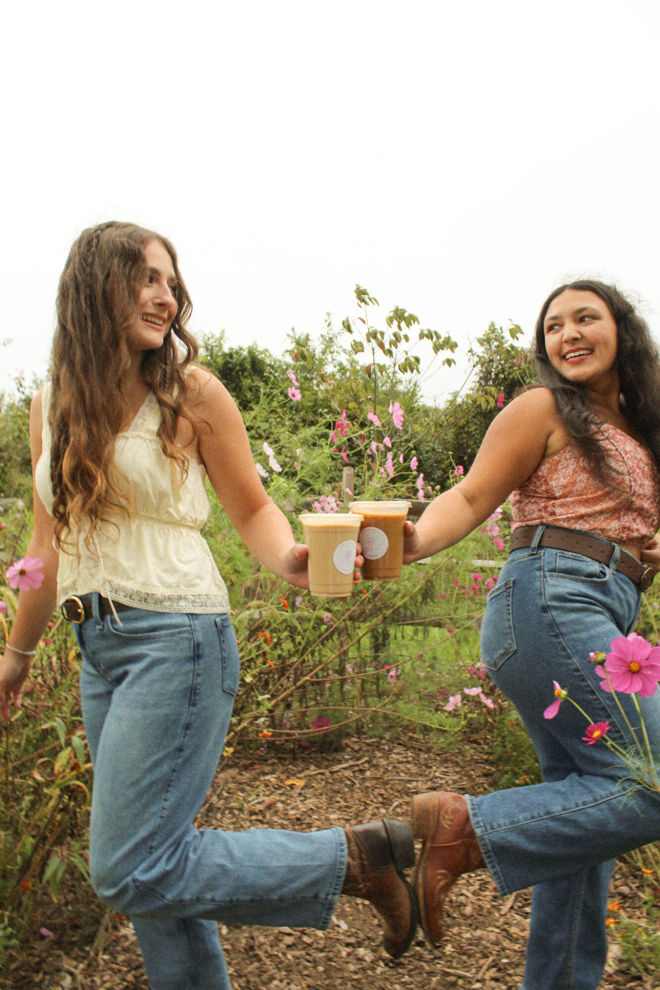 Two women holding coffee cups and smiling outdoors among flowers and greenery.