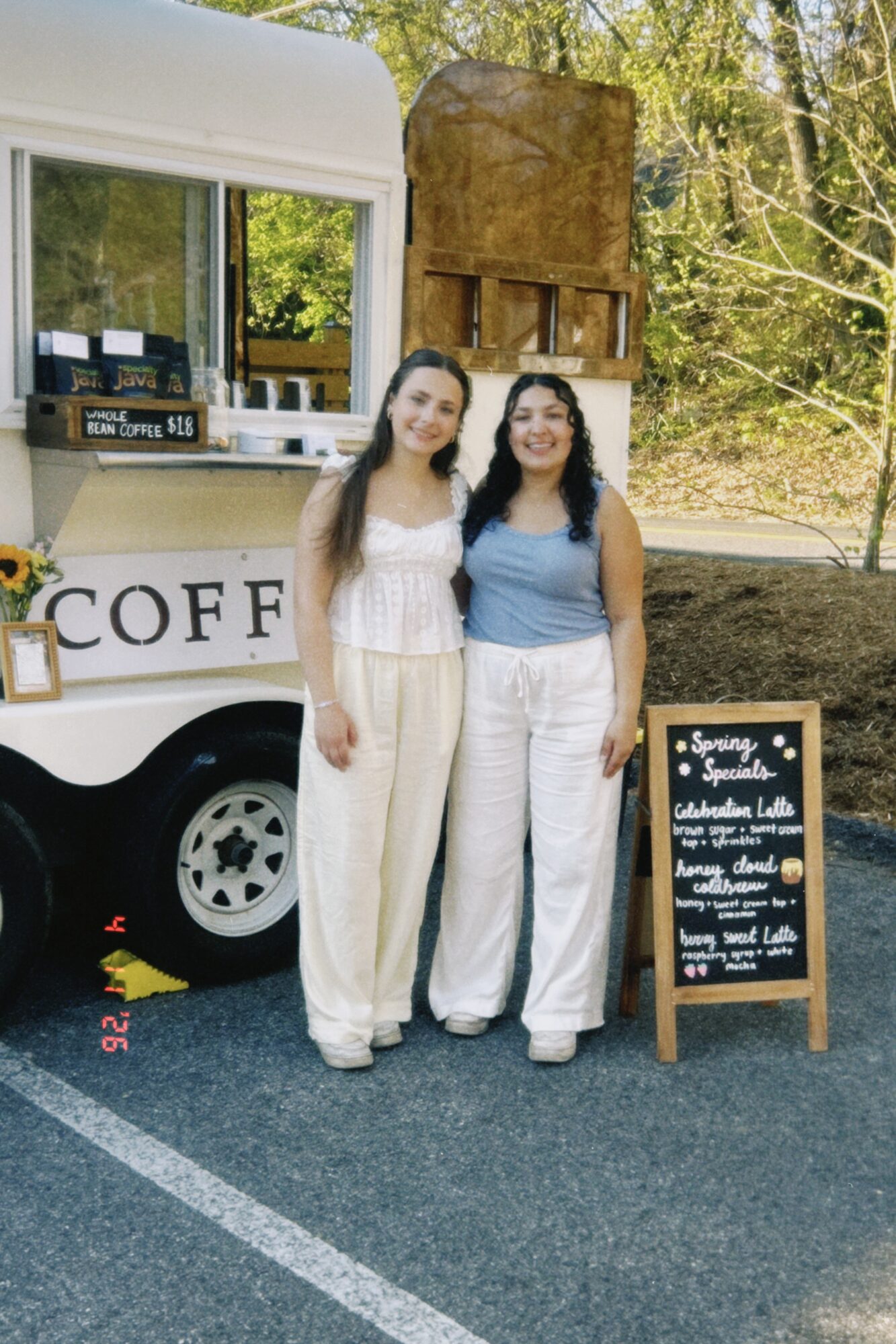 Two women stand in front of a coffee truck with a chalkboard menu, outdoors with trees in the background.