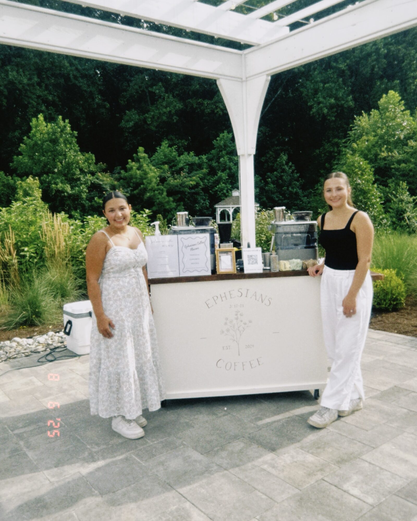 Two women stand behind a coffee stand outdoors, surrounded by greenery, under a white canopy, smiling at the camera.