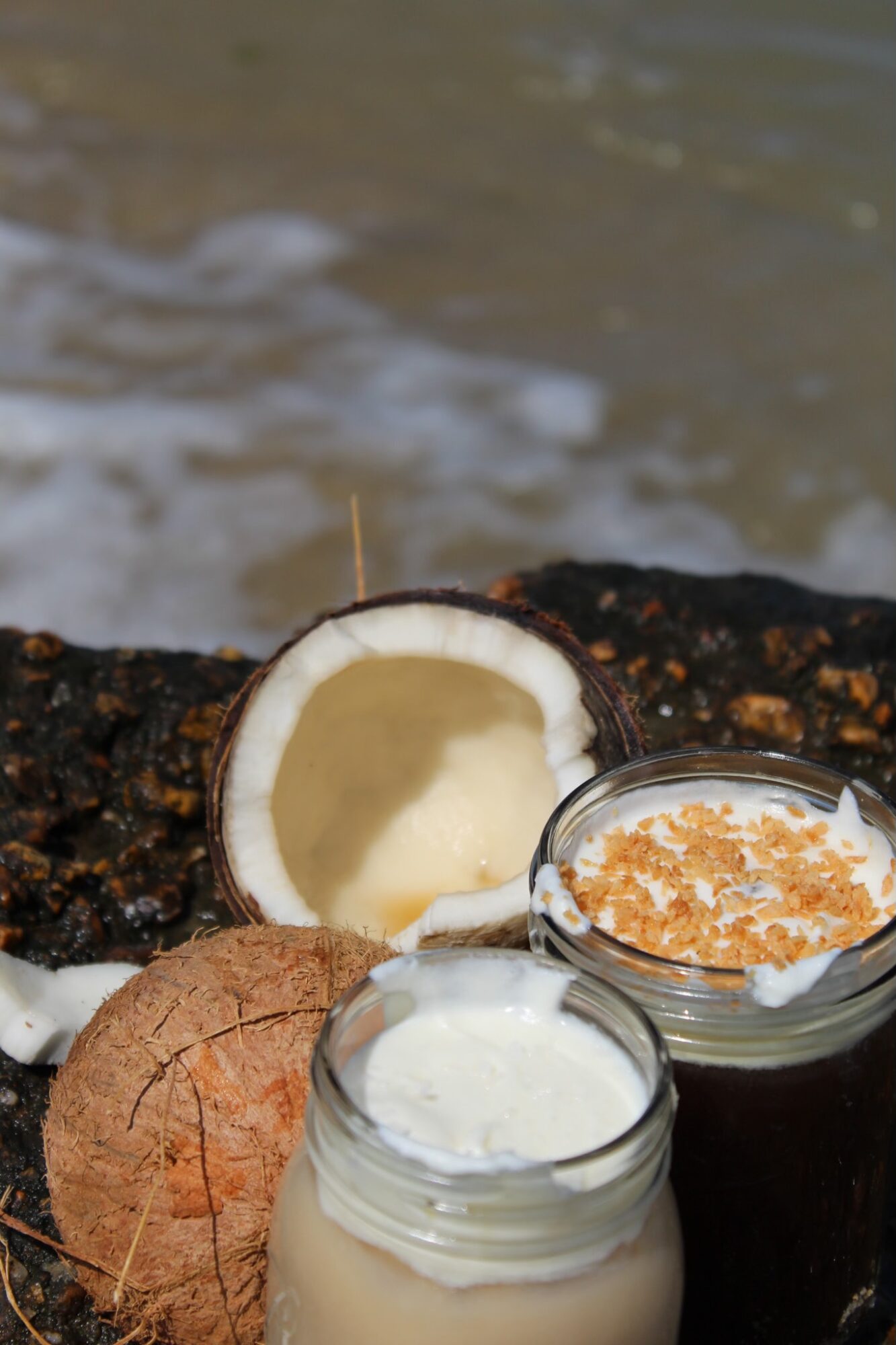 Coconut, coconut water, and a coconut shell on a rock near water, with a small bowl of brown sugar and a jar of white liquid.
